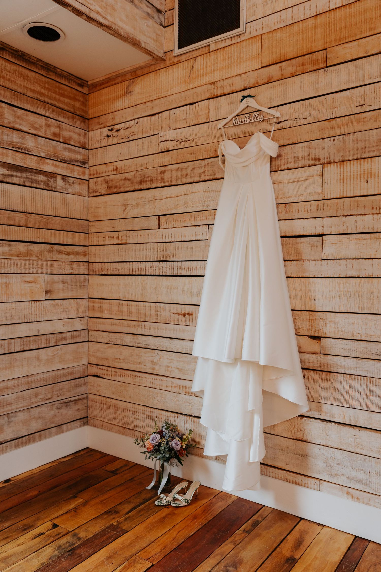 White wedding dress hanging on wooden wall with rustic decor and small floral bouquet.
