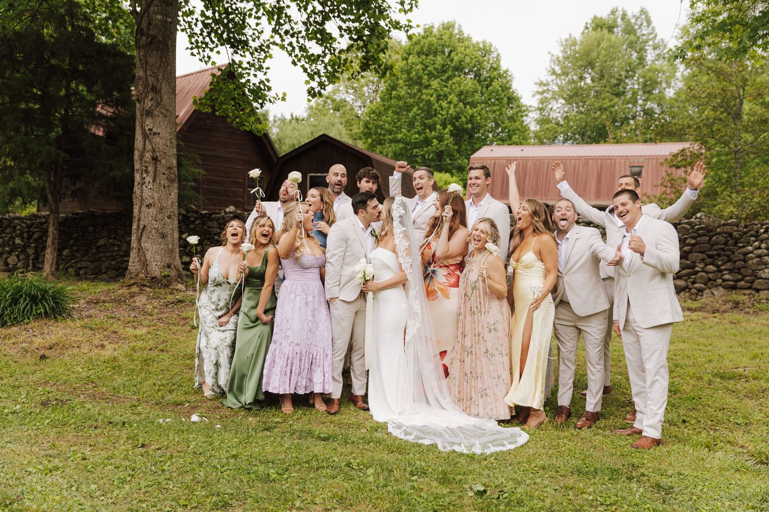 Wedding party poses together outdoors in light colored attire against stone wall backdrop.