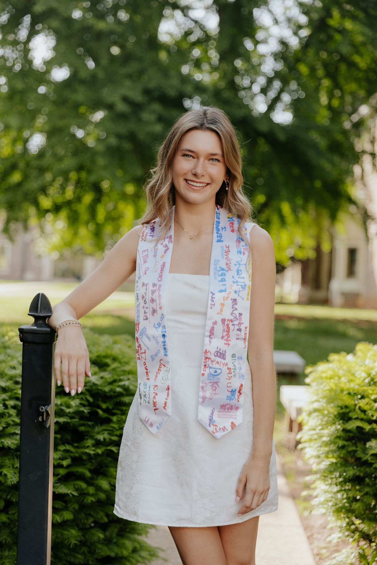 A person in a white summer dress and colorful patterned scarf stands outdoors on a garden path near a black post.