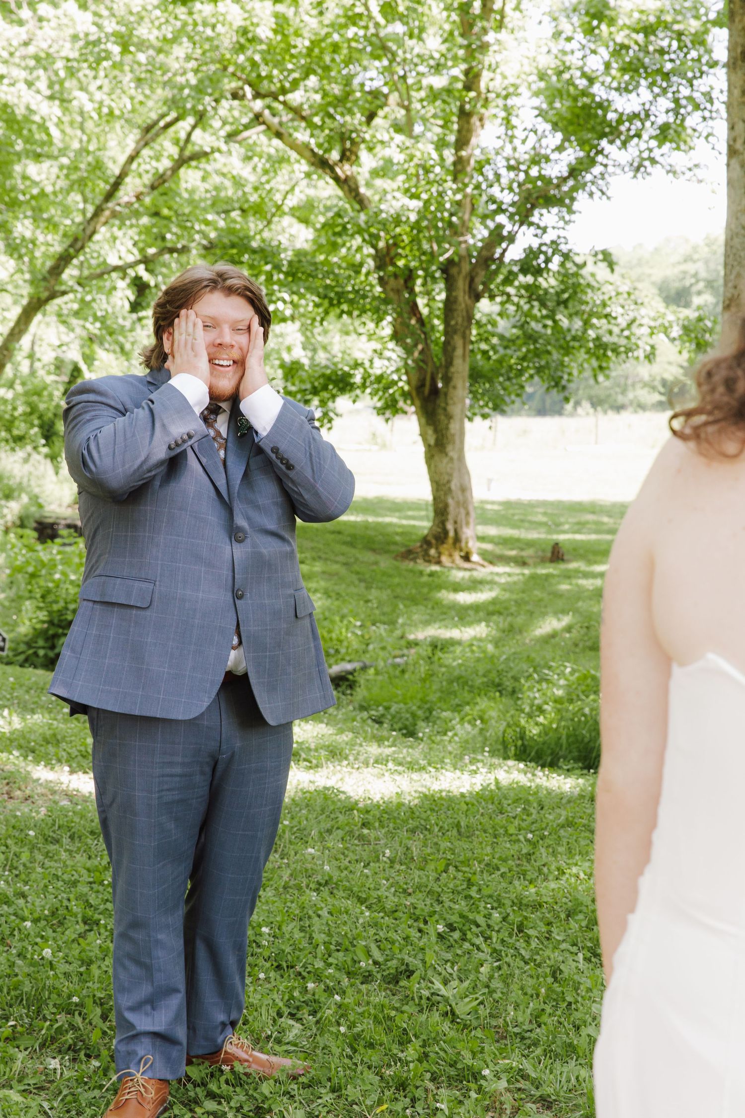 Groom covers his face with hands in emotional first look reaction seeing bride in white dress outdoors.