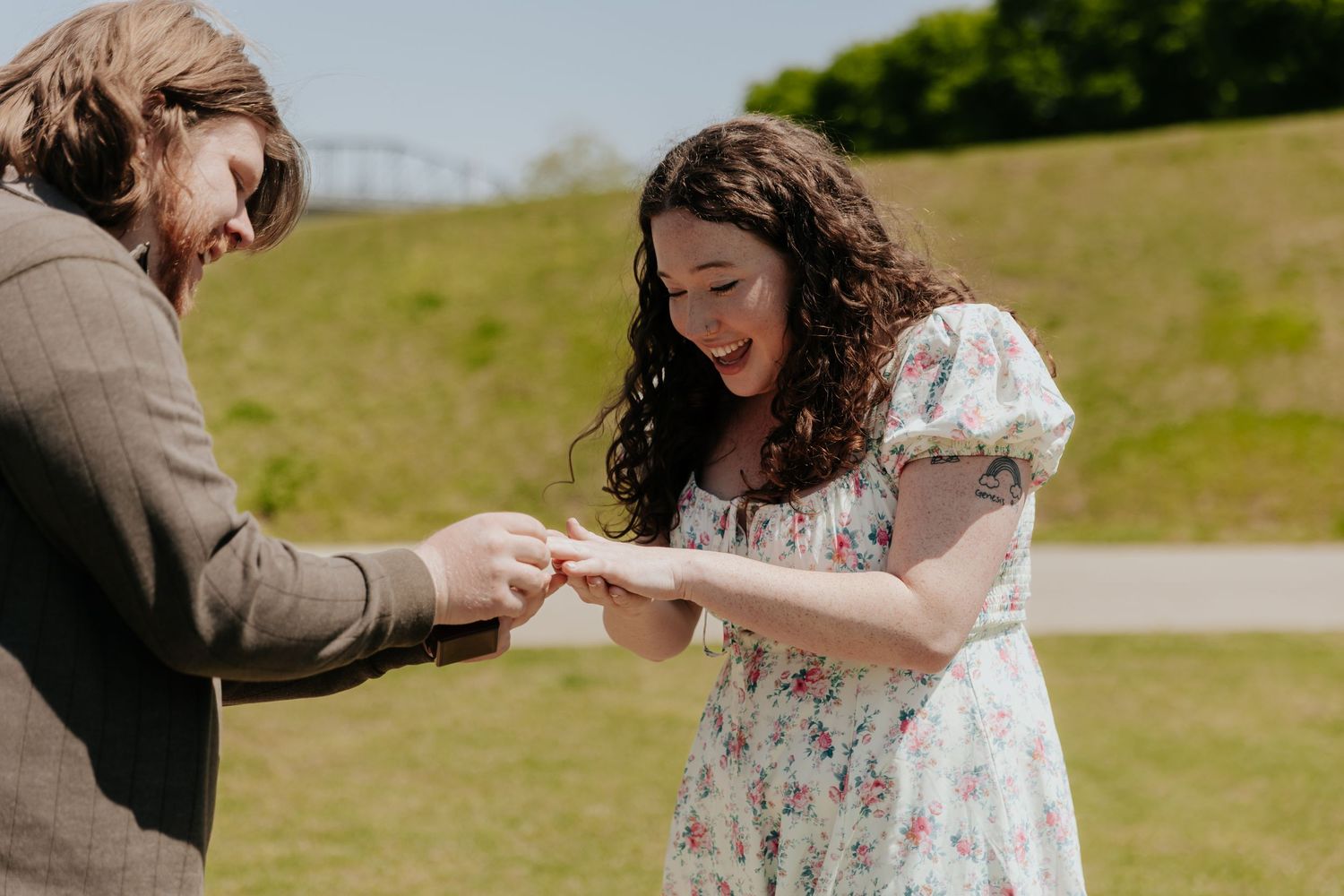 A couple shares an intimate moment during what appears to be a proposal on a grassy hill.