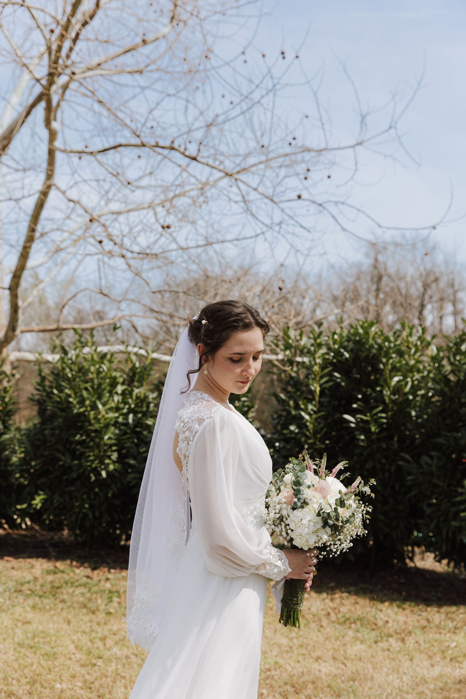 Elegant bride in white long-sleeve gown and veil holding floral bouquet in garden setting.