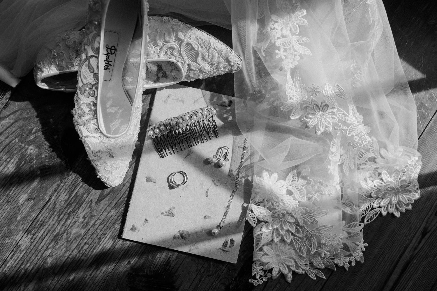 Black and white flat lay of bridal accessories including lace heels, veil, rings, and a comb on a wooden surface.