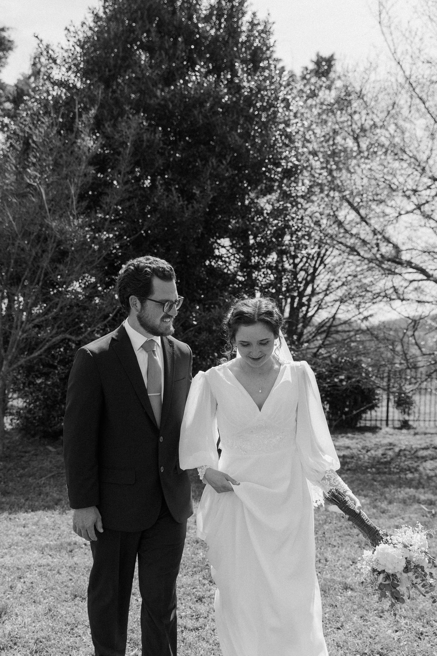 Black and white photo of bride and groom walking outdoors, bride holding bouquet in elegant long-sleeve wedding dress.