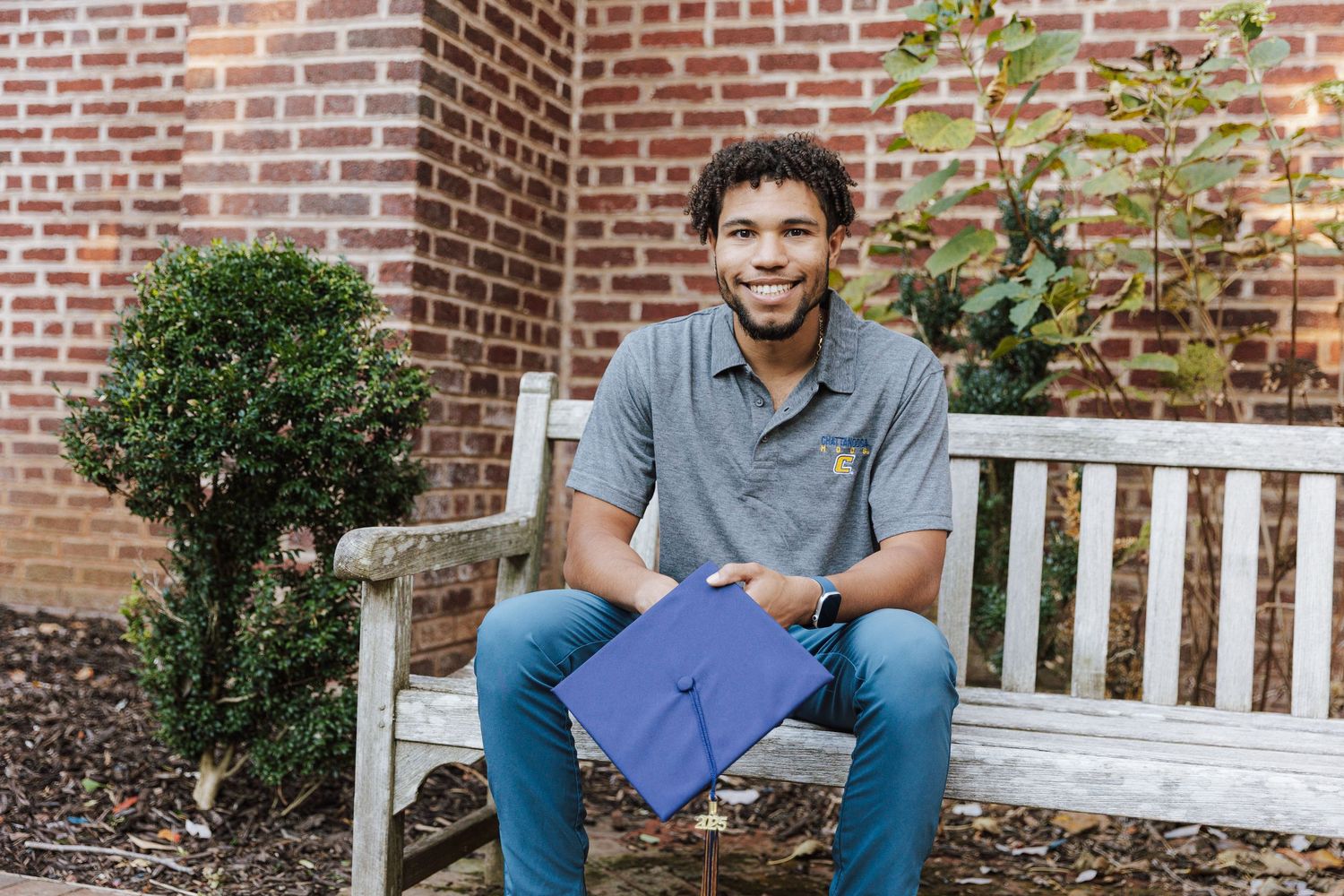Graduate in blue regalia sits on white bench holding diploma near brick building.