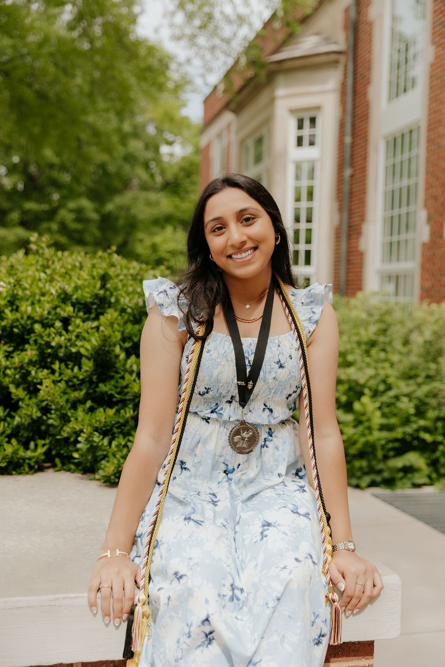 Graduate in a floral dress and medal poses outdoors near brick buildings.