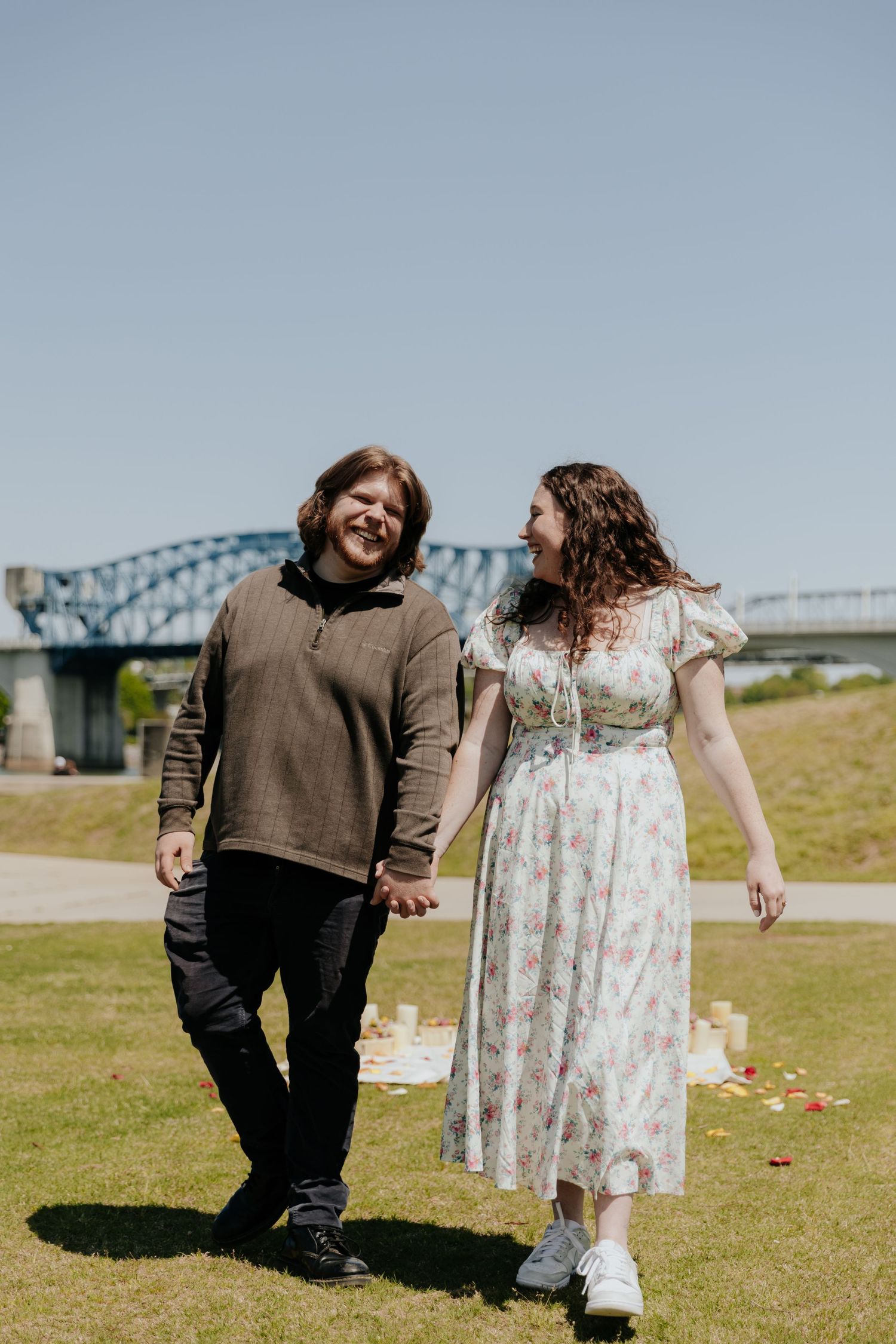 A couple walking hand in hand on a sunny day with a bridge visible in the distance.