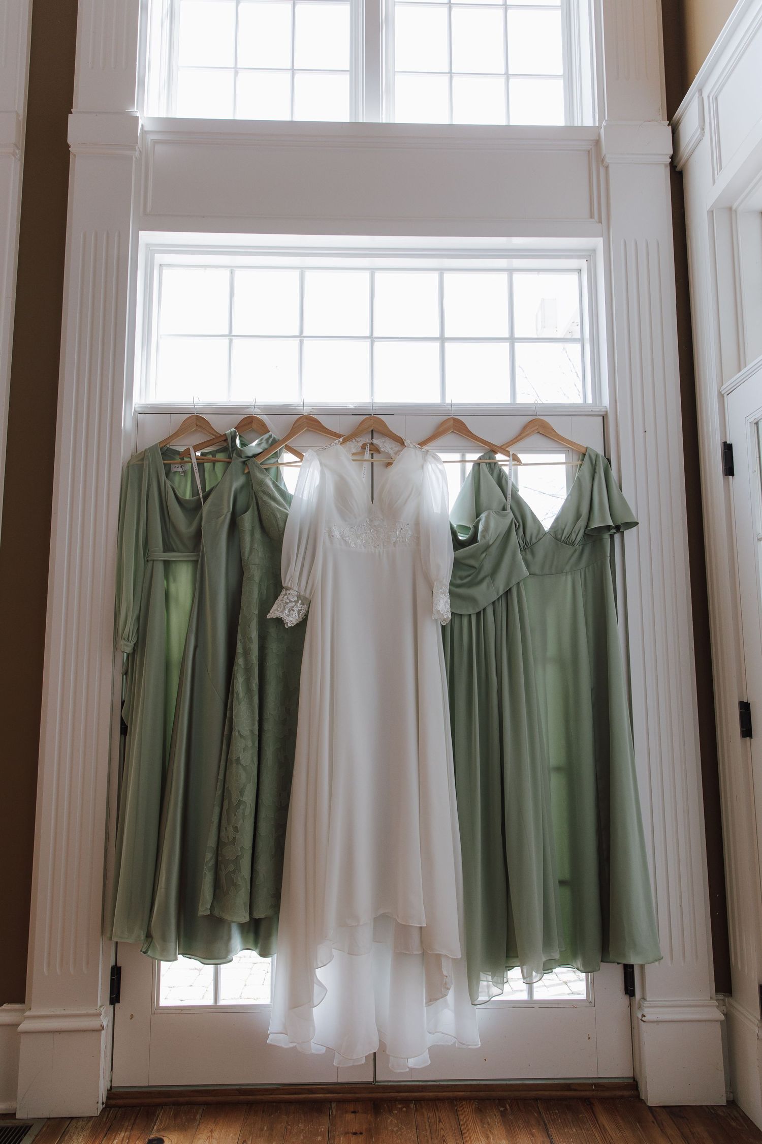 Wedding and bridesmaid dresses in white and sage green hanging against bright windows.