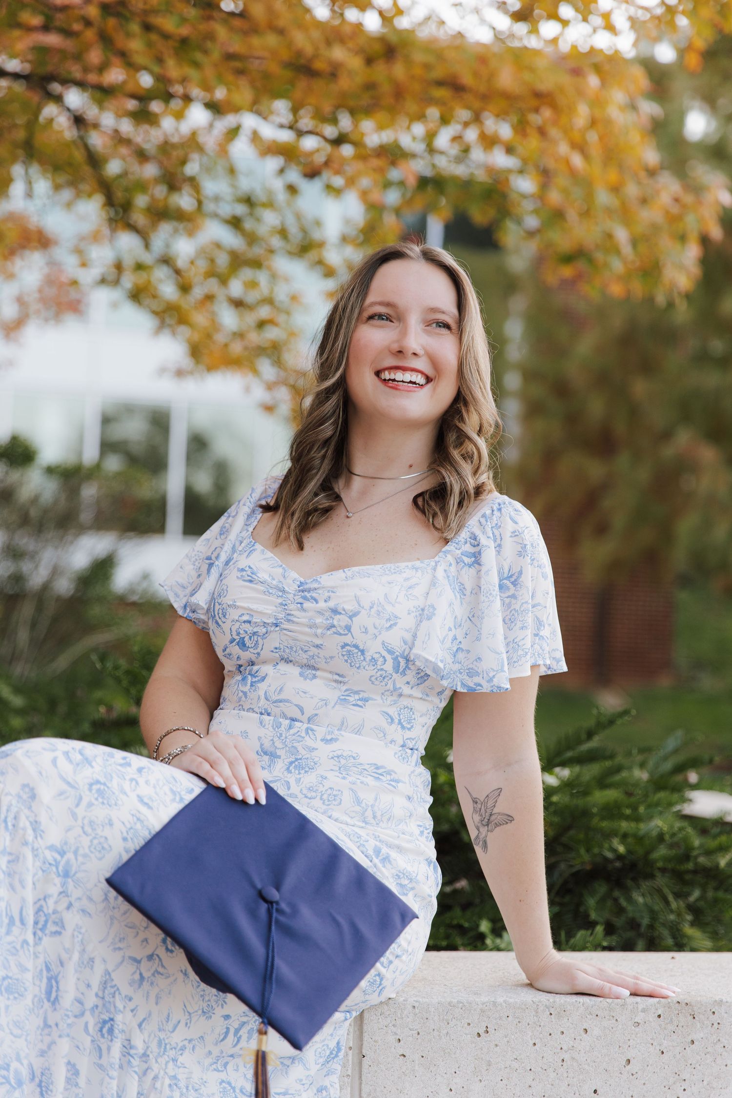 Graduate holding blue diploma folder poses on white stone ledge surrounded by fall foliage.
