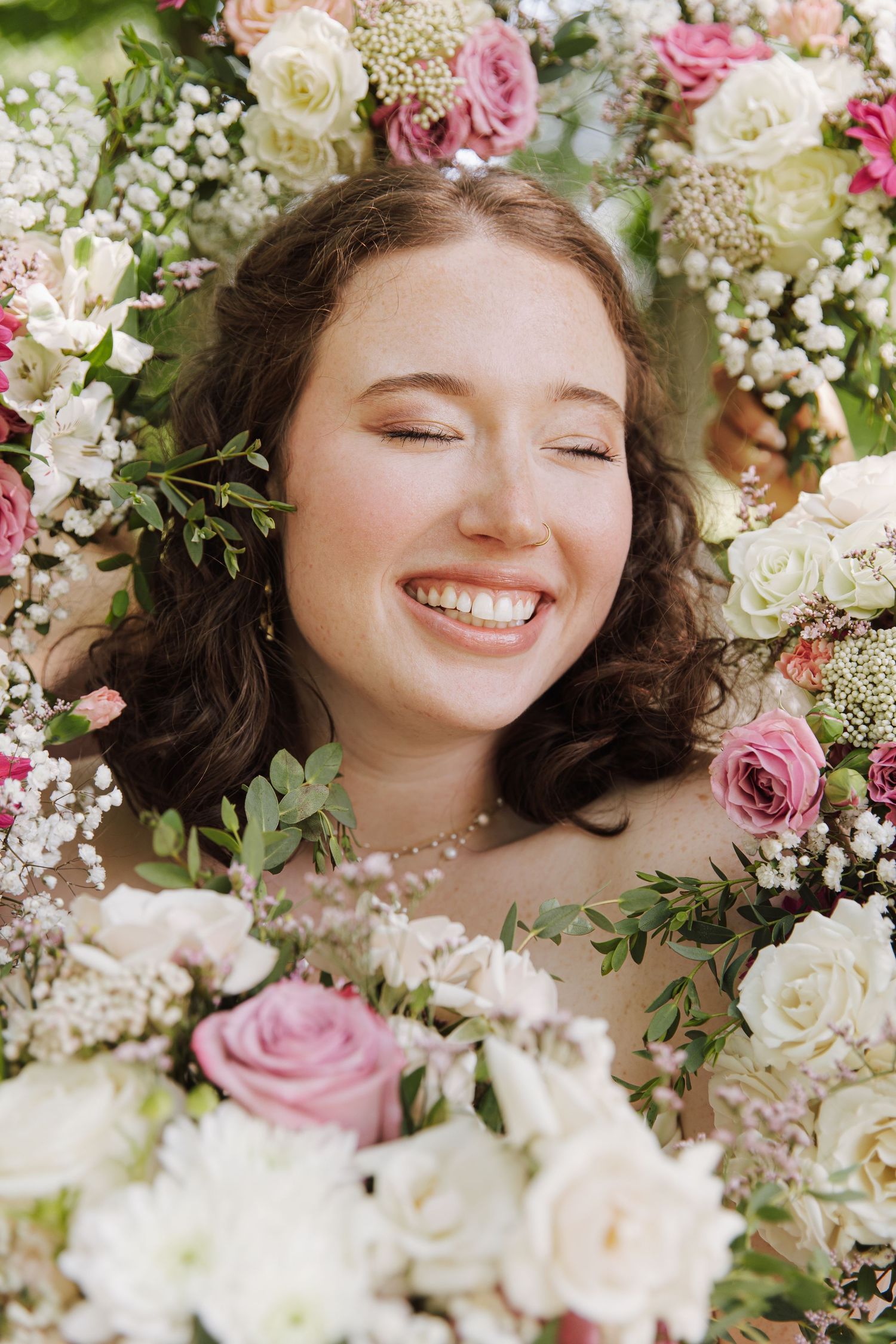 Smiling young woman surrounded by pink and white roses, baby's breath, and greenery in a floral portrait.