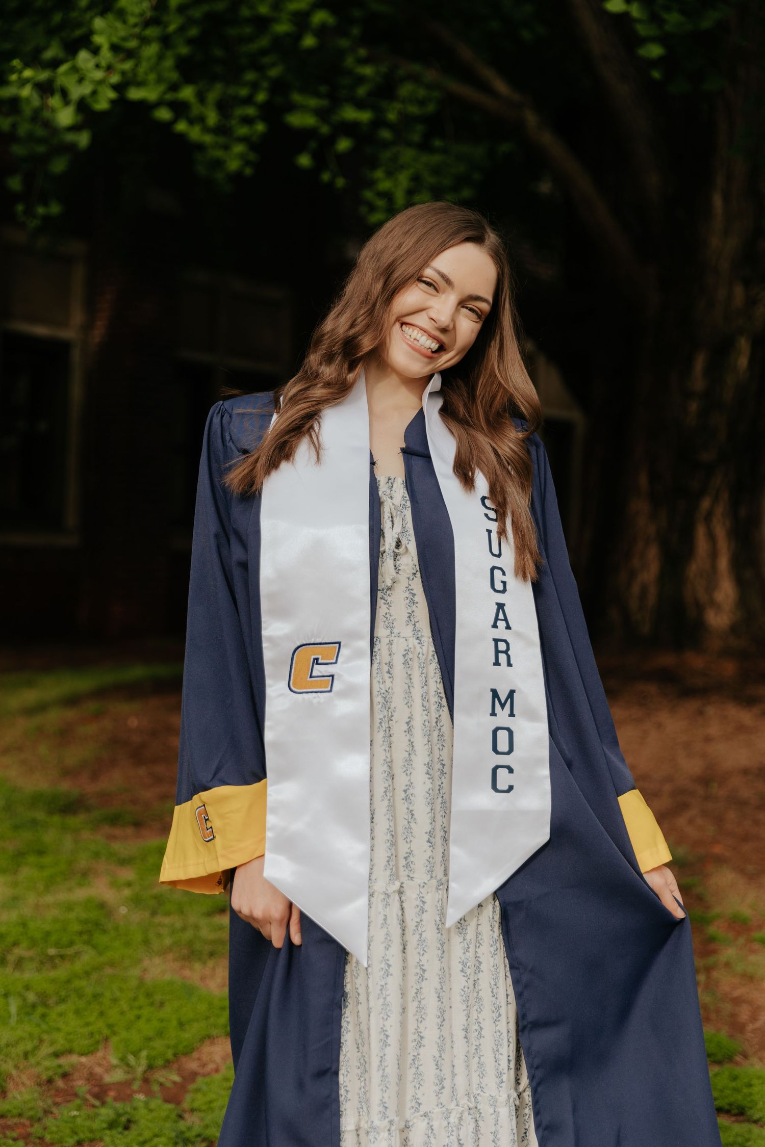 A graduate in a navy blue and gold cap and gown poses outdoors against a wooded background.