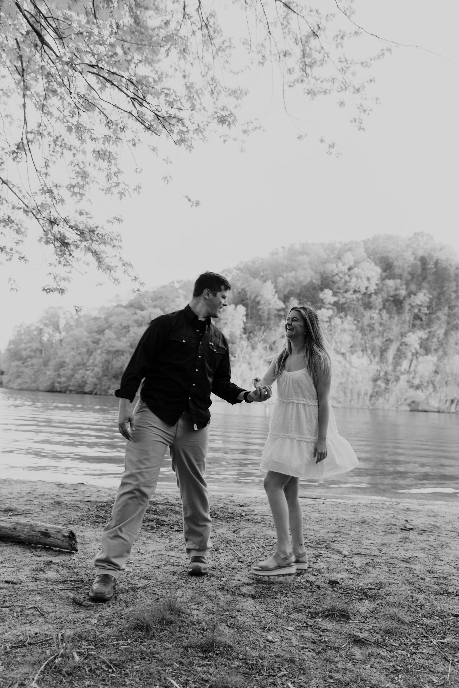 A couple holds hands on a riverbank with cliffs and trees creating a scenic backdrop.