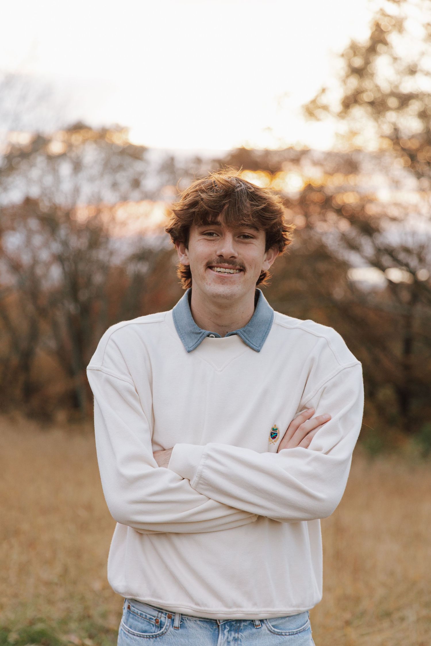 Casual portrait of person in white sweater and jeans standing in autumn field at sunset.