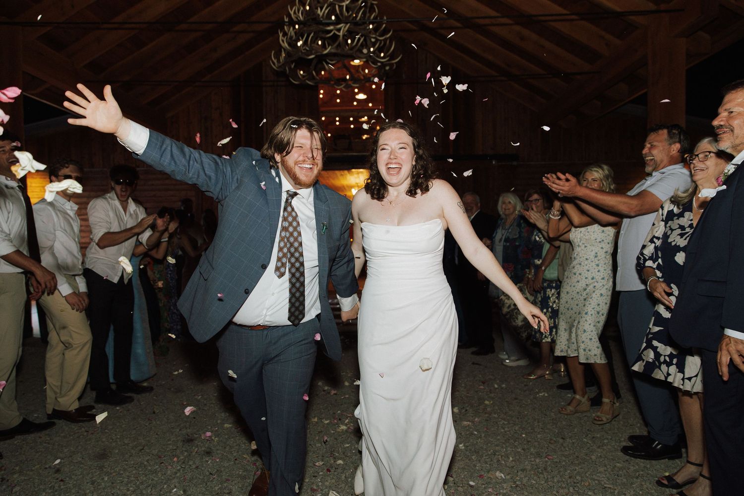Joyful newlyweds exit wedding reception as guests toss flower petals in rustic barn venue.