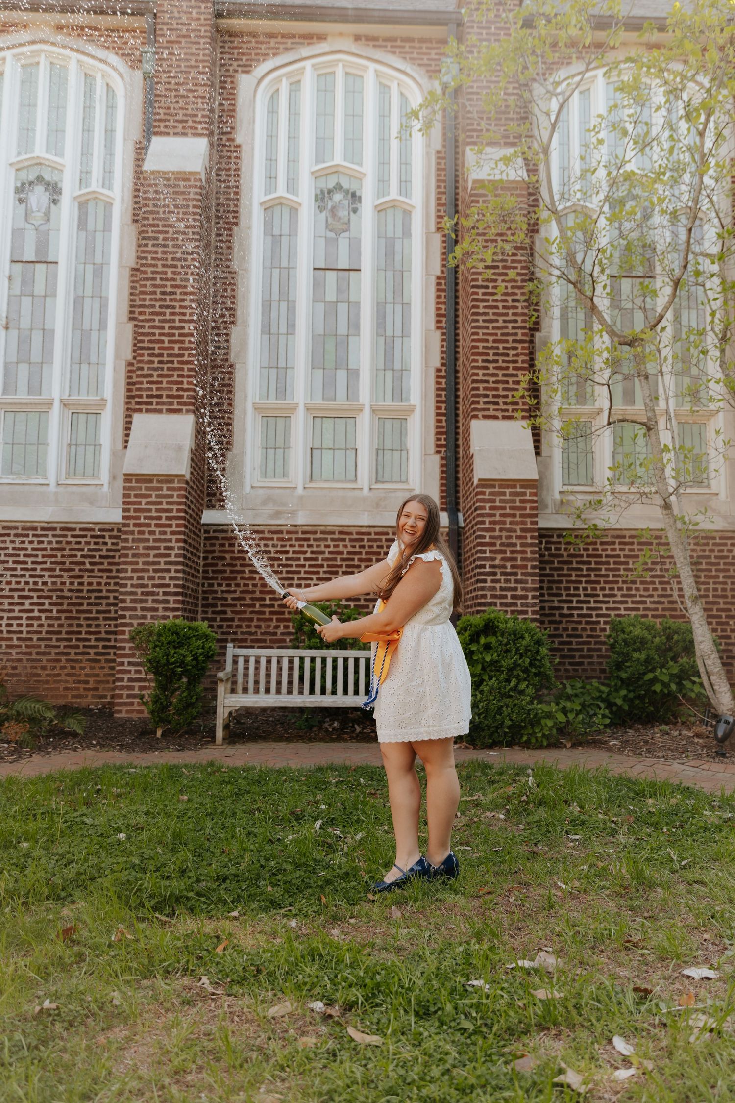 A person in a white dress poses playfully in front of a brick building with arched windows.
