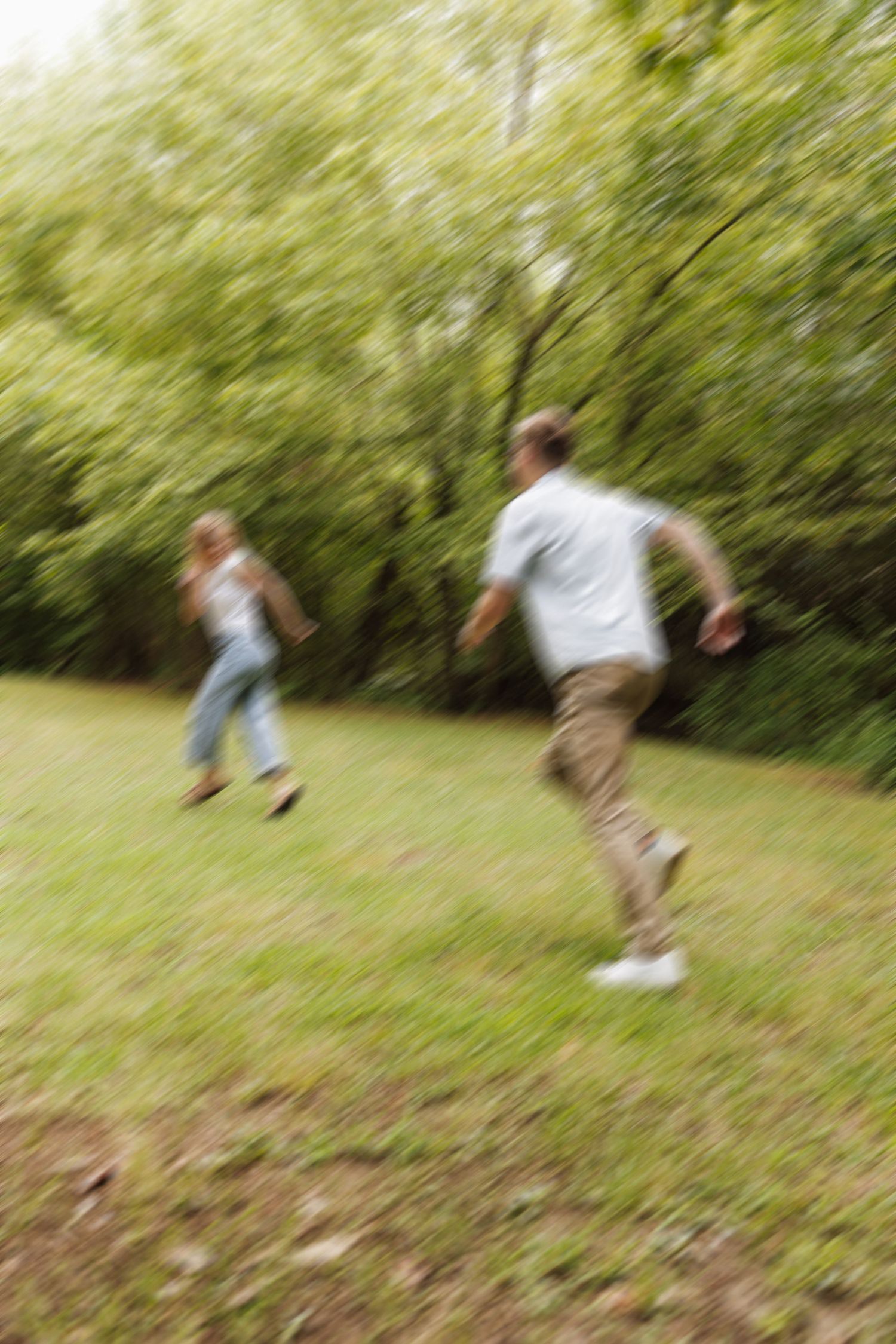 Two people playfully run through a grassy area with trees in the background.