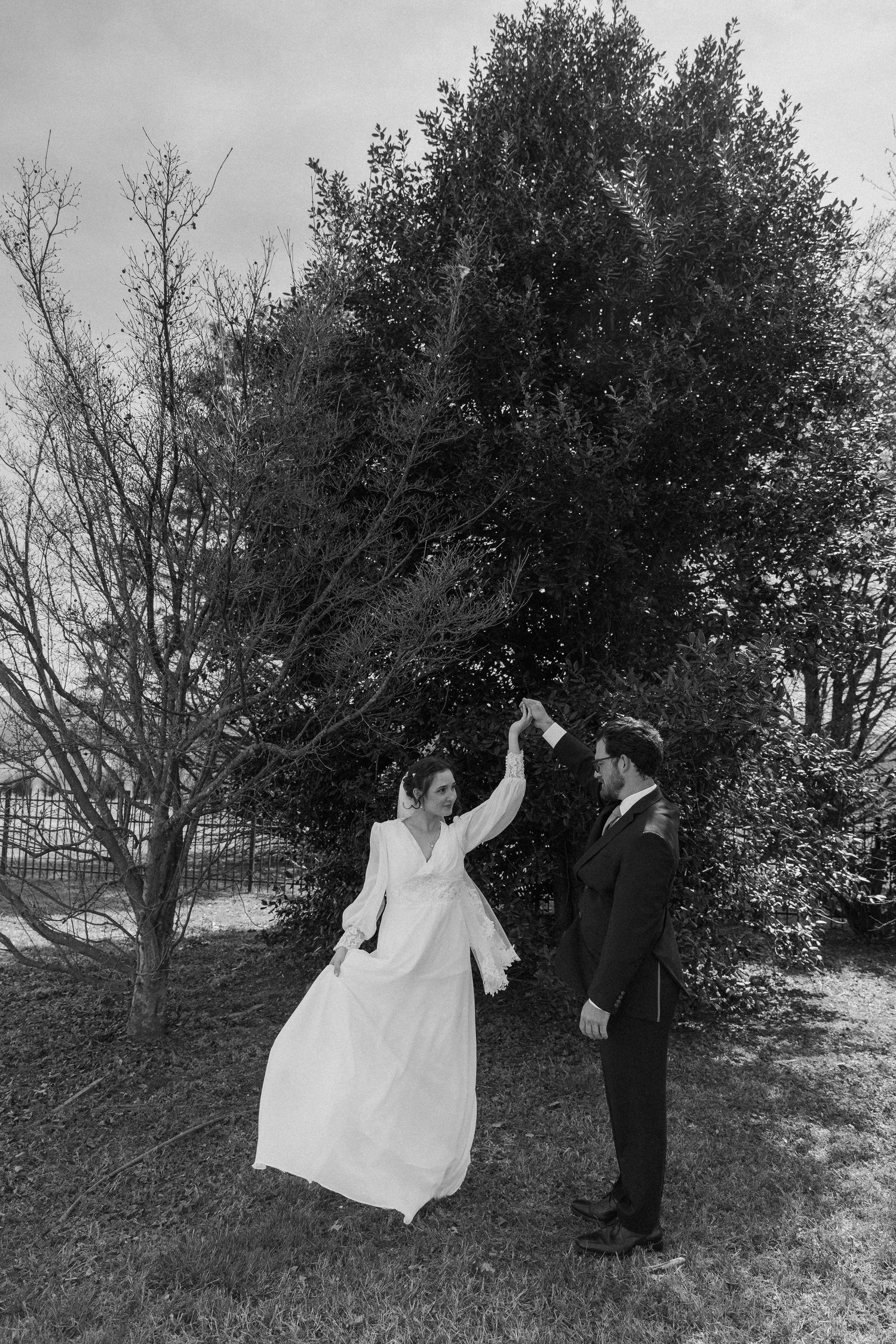 Black and white photo of groom spinning bride in white gown outdoors near trees on wedding day.
