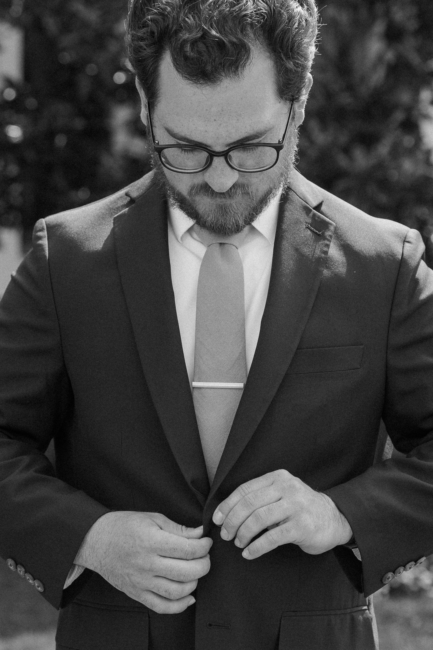 Black and white photo of bearded man in suit adjusting jacket buttons outdoors.