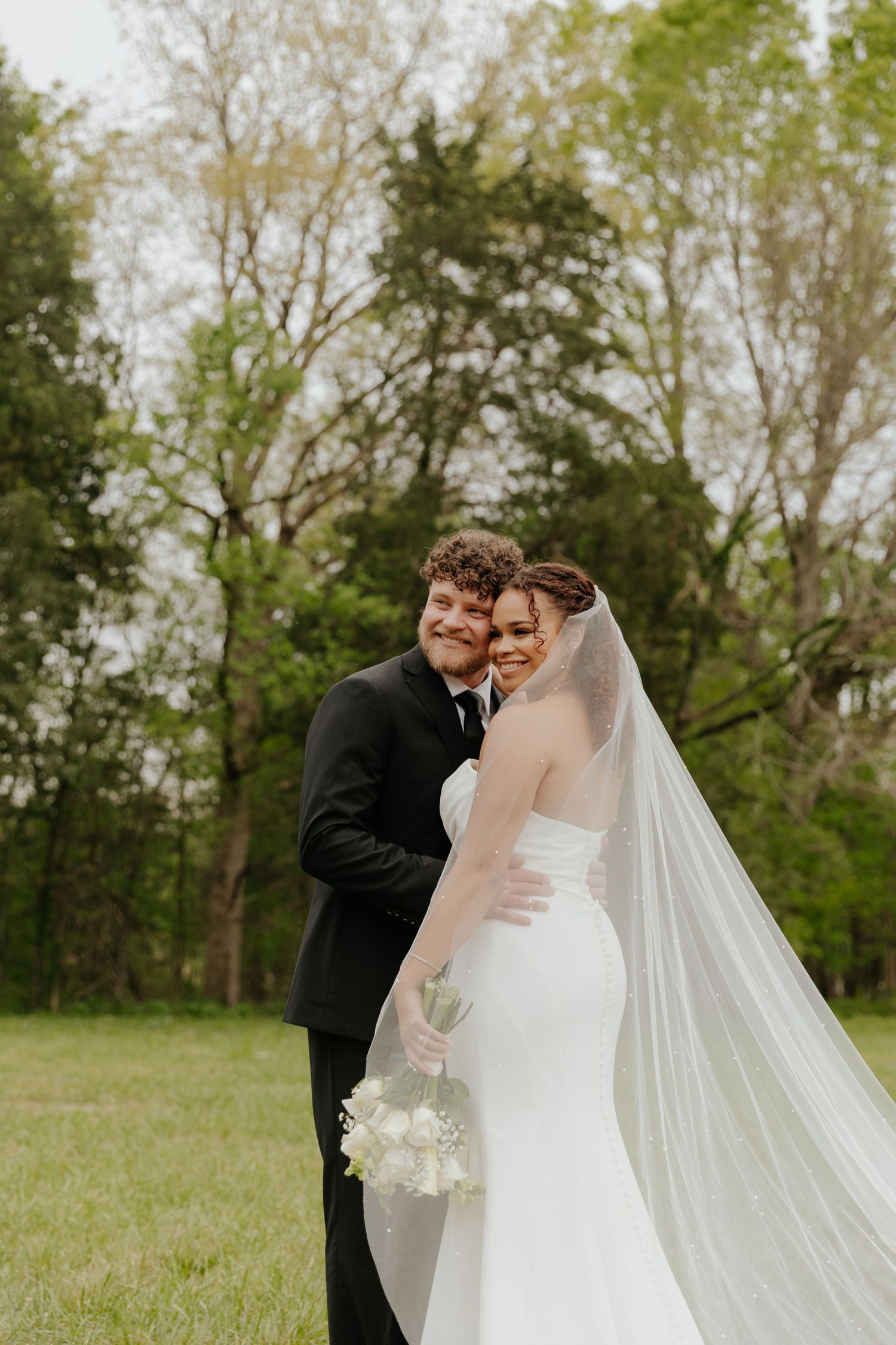 Couple in wedding attire embracing in a green field with flowing veil.