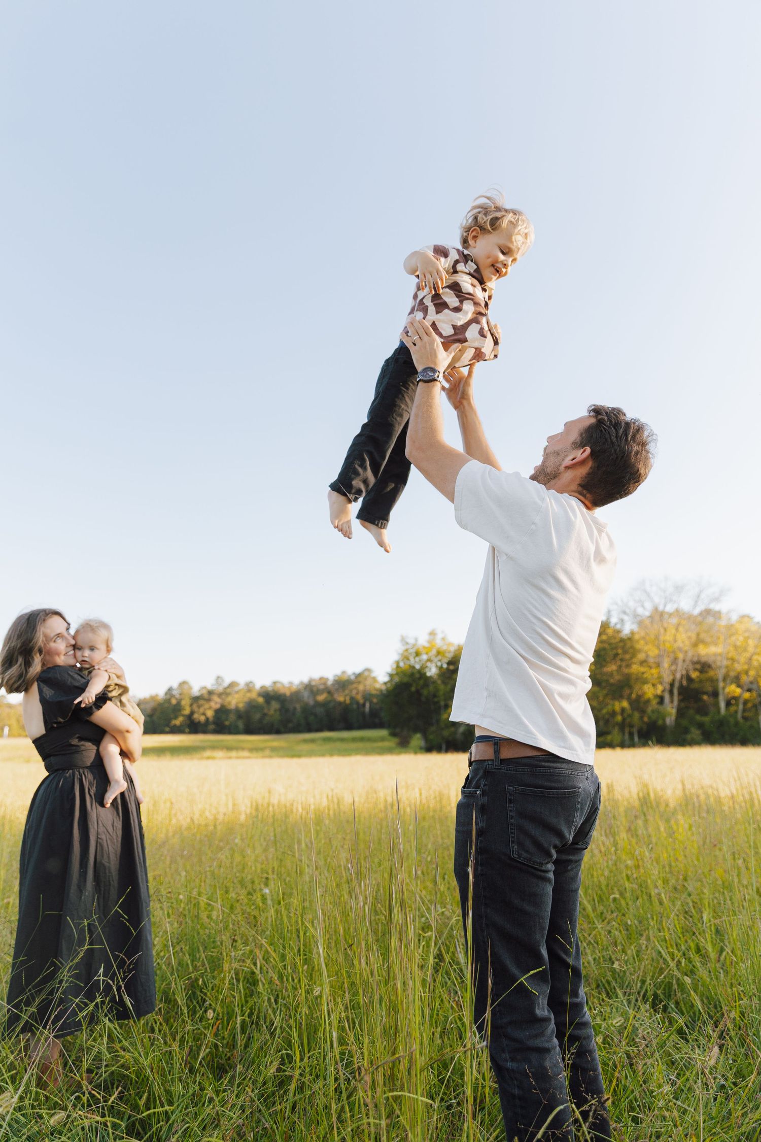 Family enjoying a playful moment together in a sunny meadow with tall grass and trees in the background.