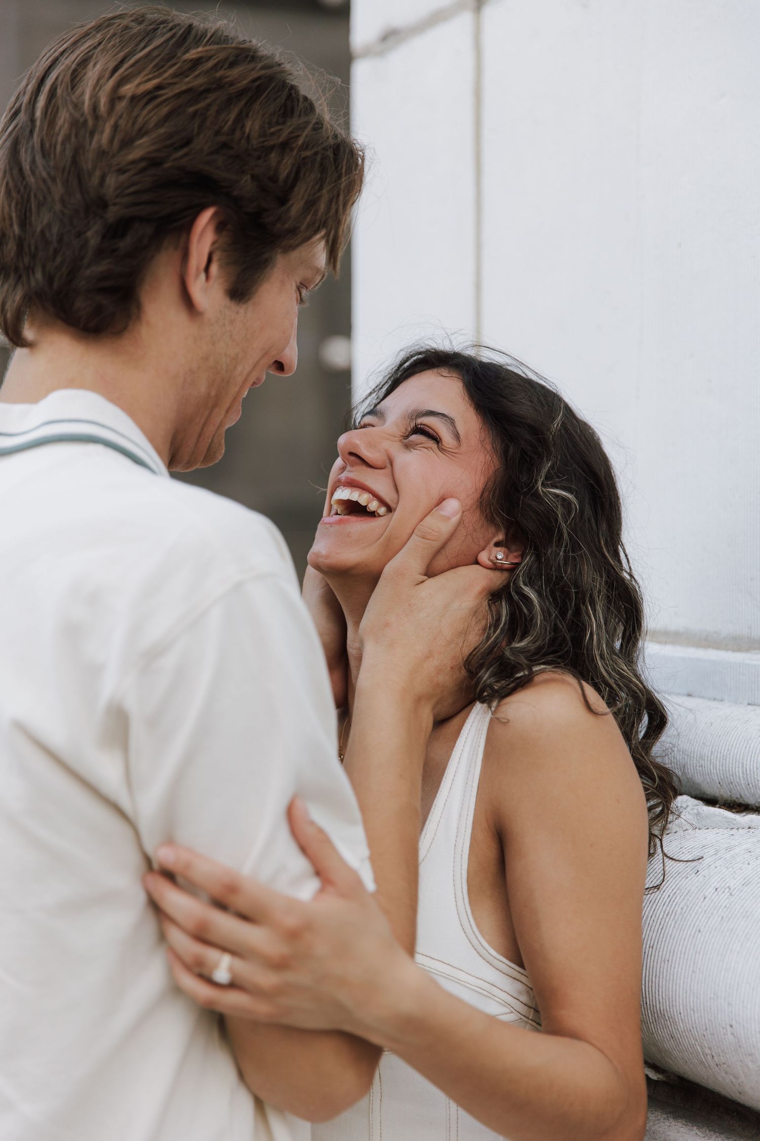 Joyful bride in white dress laughing with groom during romantic outdoor engagement photo session.