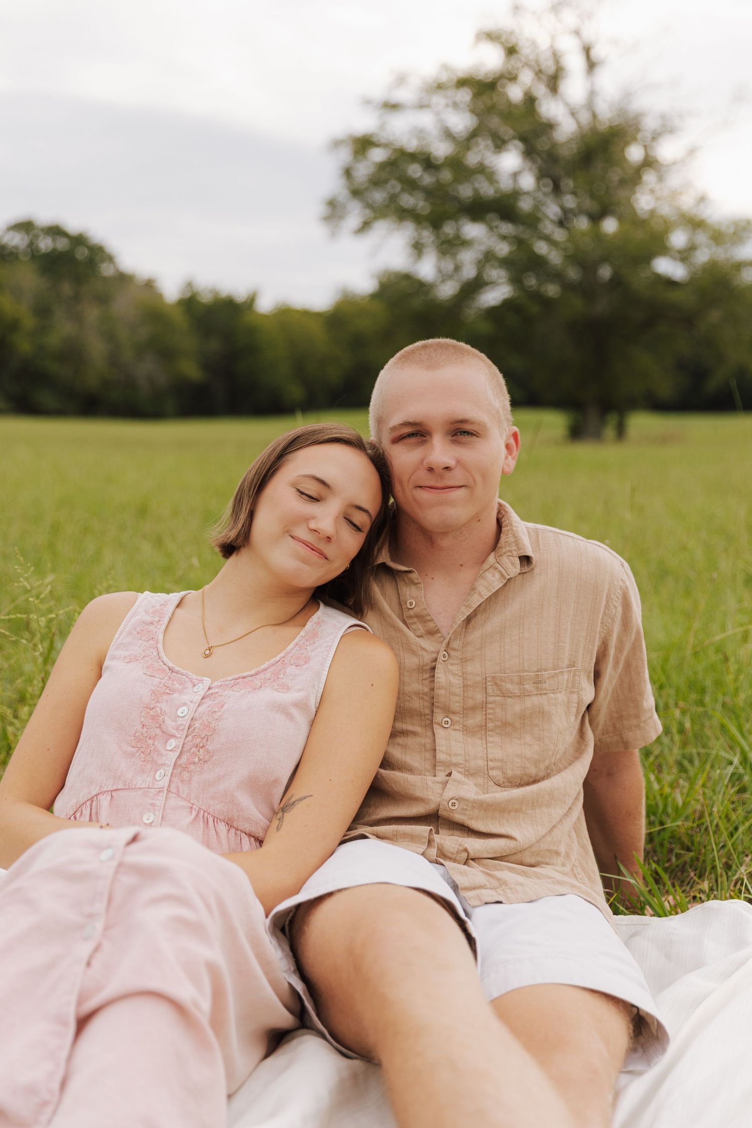 A couple relaxes together on a blanket in a sunny meadow wearing summer attire.