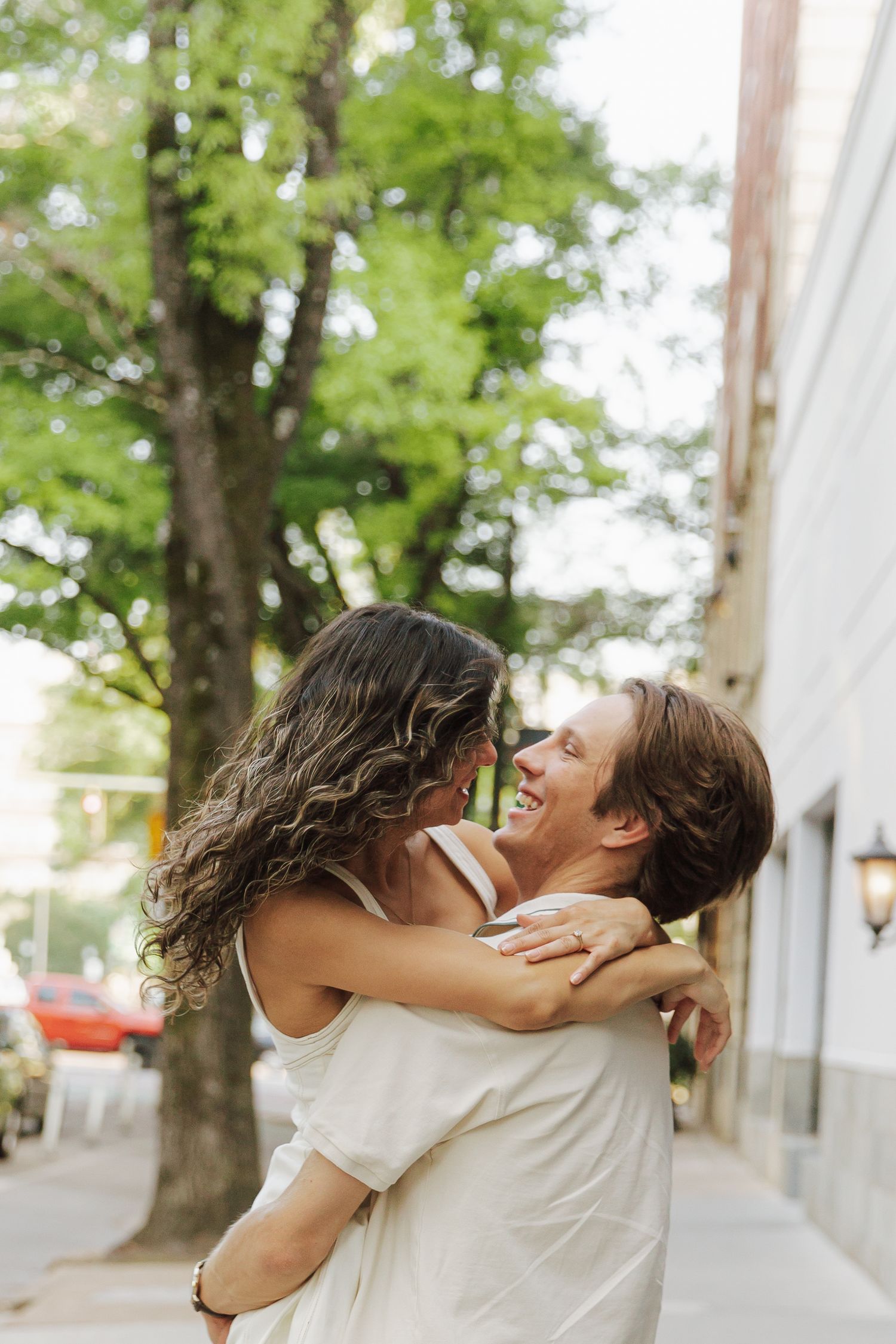 A joyful couple embraces and laughs together on a sunny urban sidewalk surrounded by lush green trees.