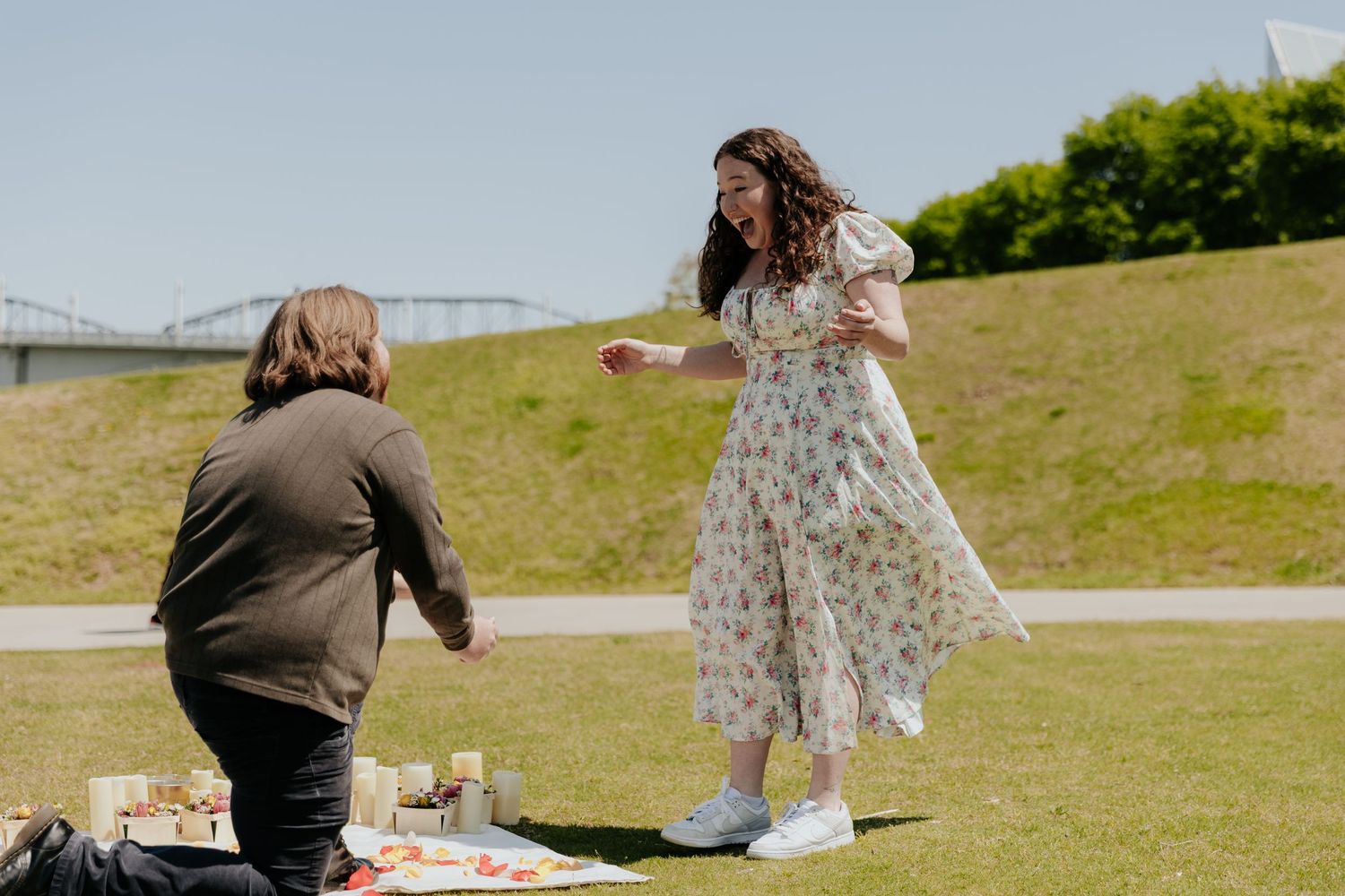 A proposal scene unfolds on a grassy hill with a bridge visible in the background.
