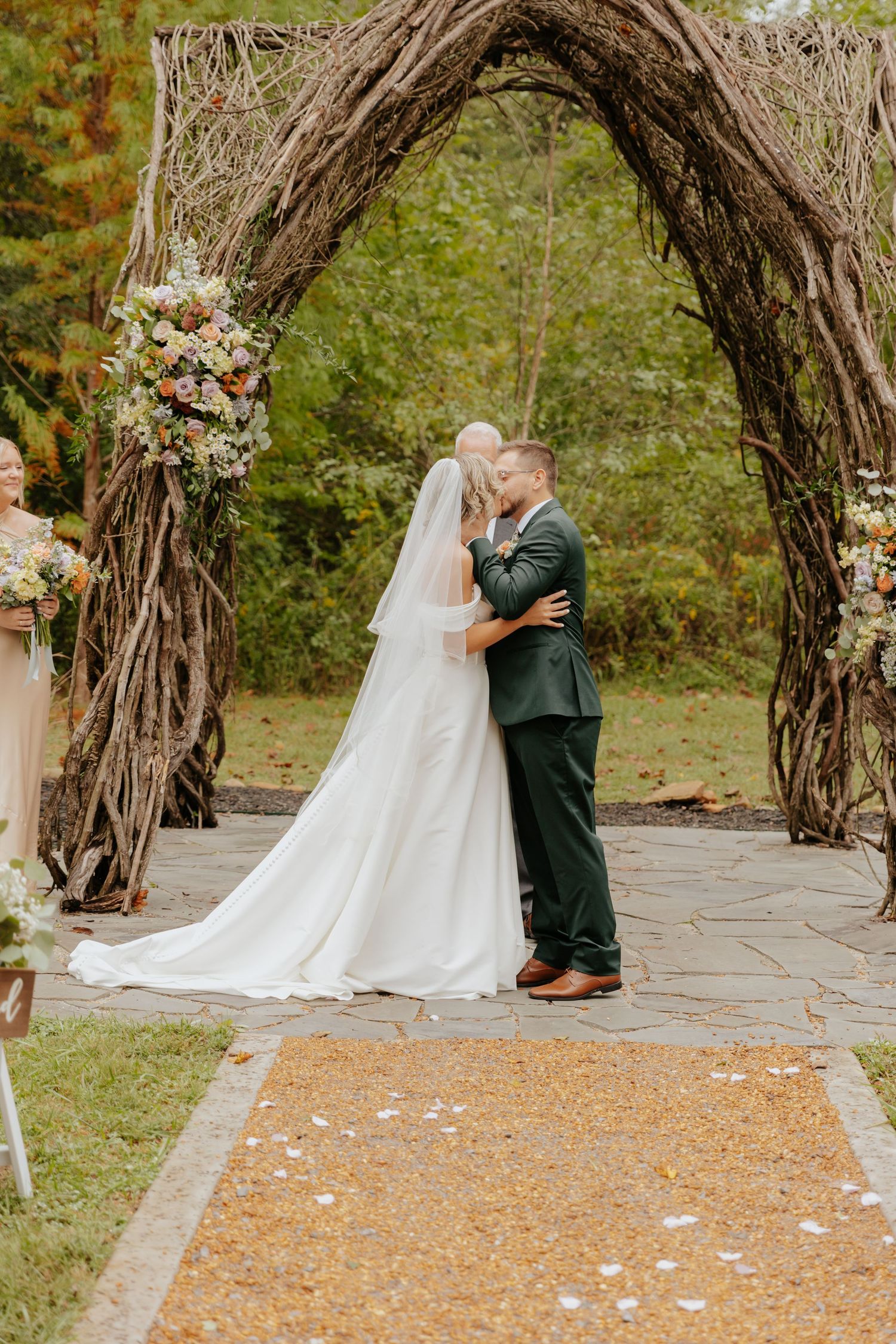 Wedding ceremony under rustic wooden arch with rose petals scattered on stone path.