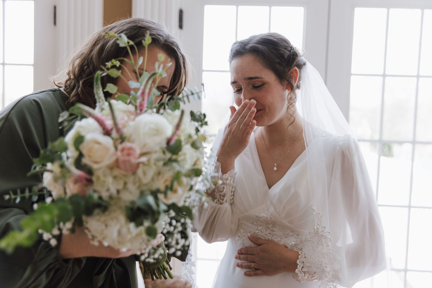 Emotional bride in white veil touches her face while bridesmaid holds a beautiful bouquet of white and pink flowers.