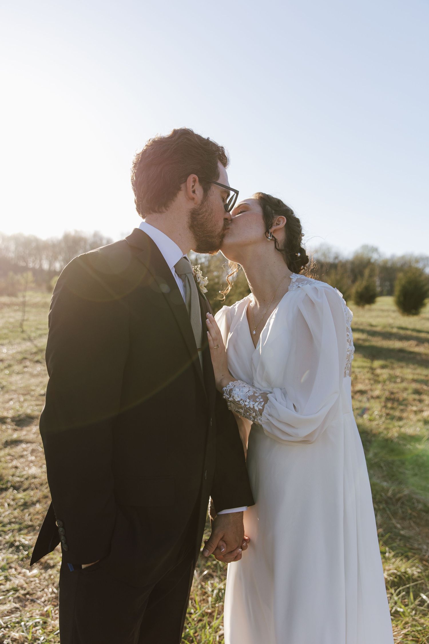 Wedding couple kissing in sunny field, groom in dark suit with tie, bride in elegant long white dress holding hands.