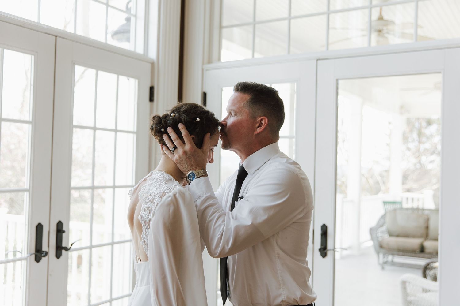 Emotional father kissing bride on forehead near white French doors before wedding ceremony.