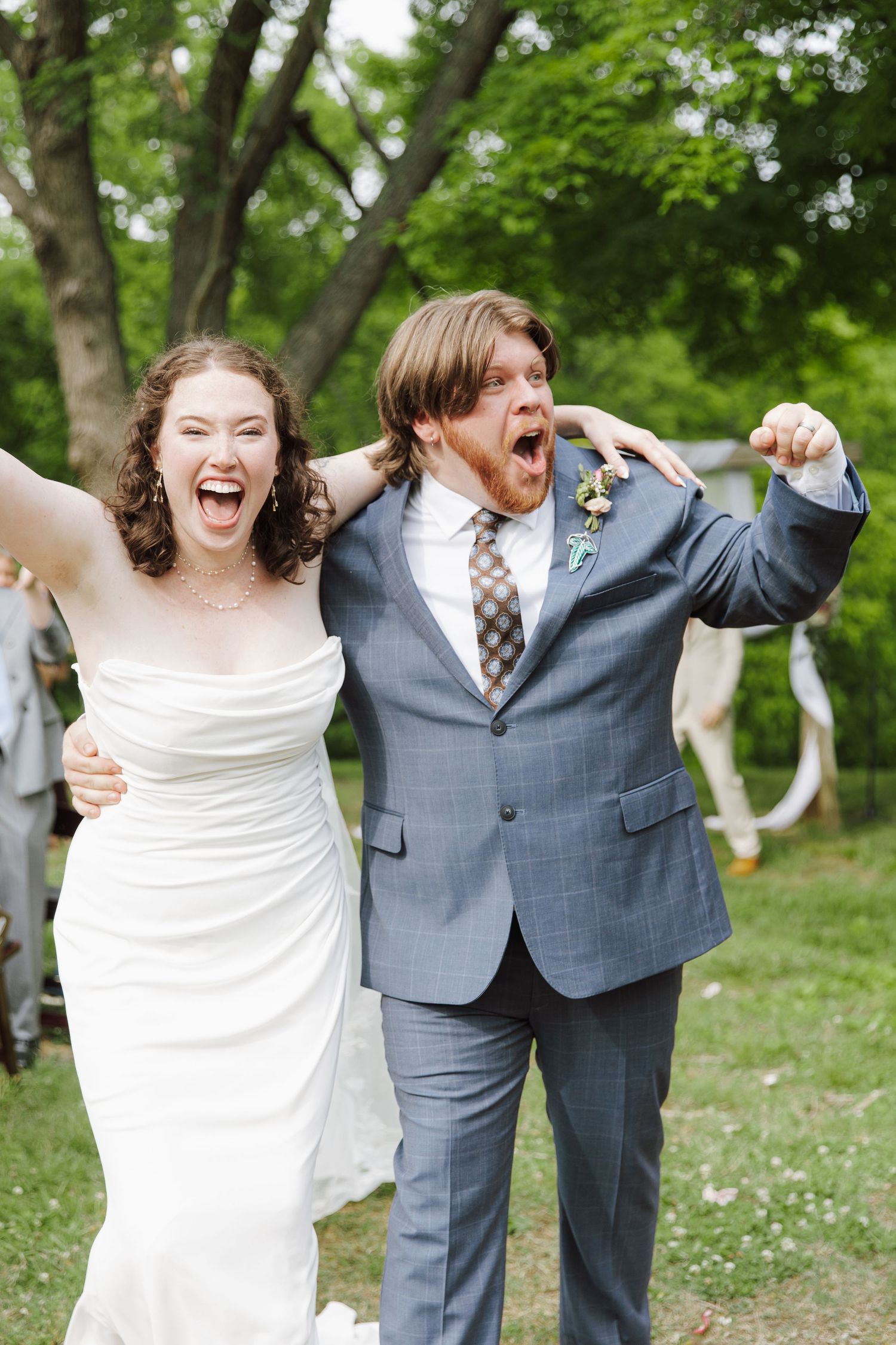 Joyful newlyweds celebrate outdoors, bride in white gown and groom in gray suit cheering with arms raised.