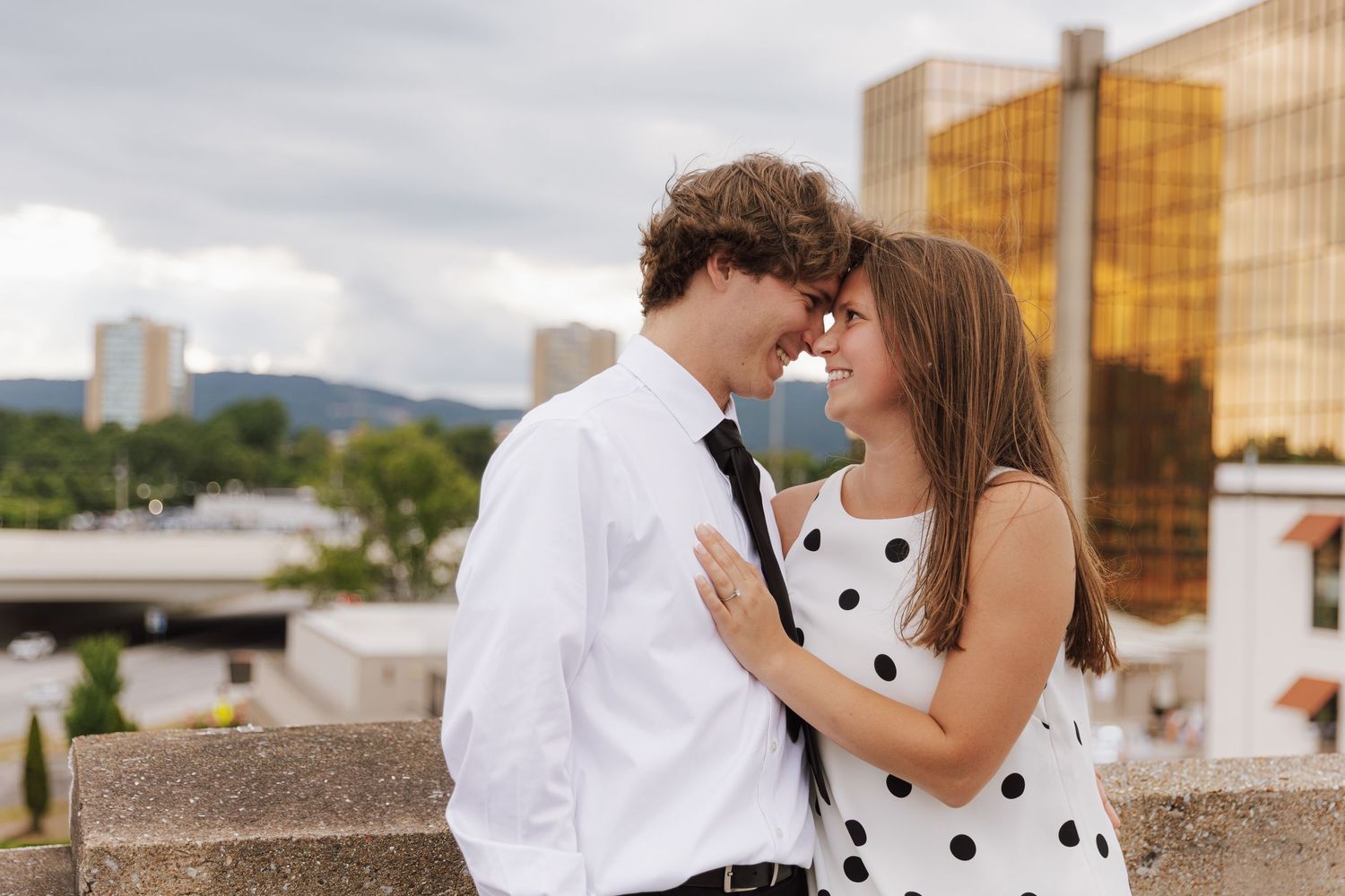 A couple in formal attire poses against an urban backdrop with golden buildings.
