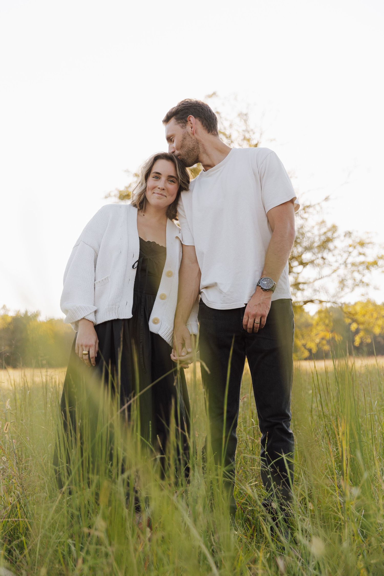 A couple stands together in a sunlit field of tall grass wearing casual white outfits during golden hour.