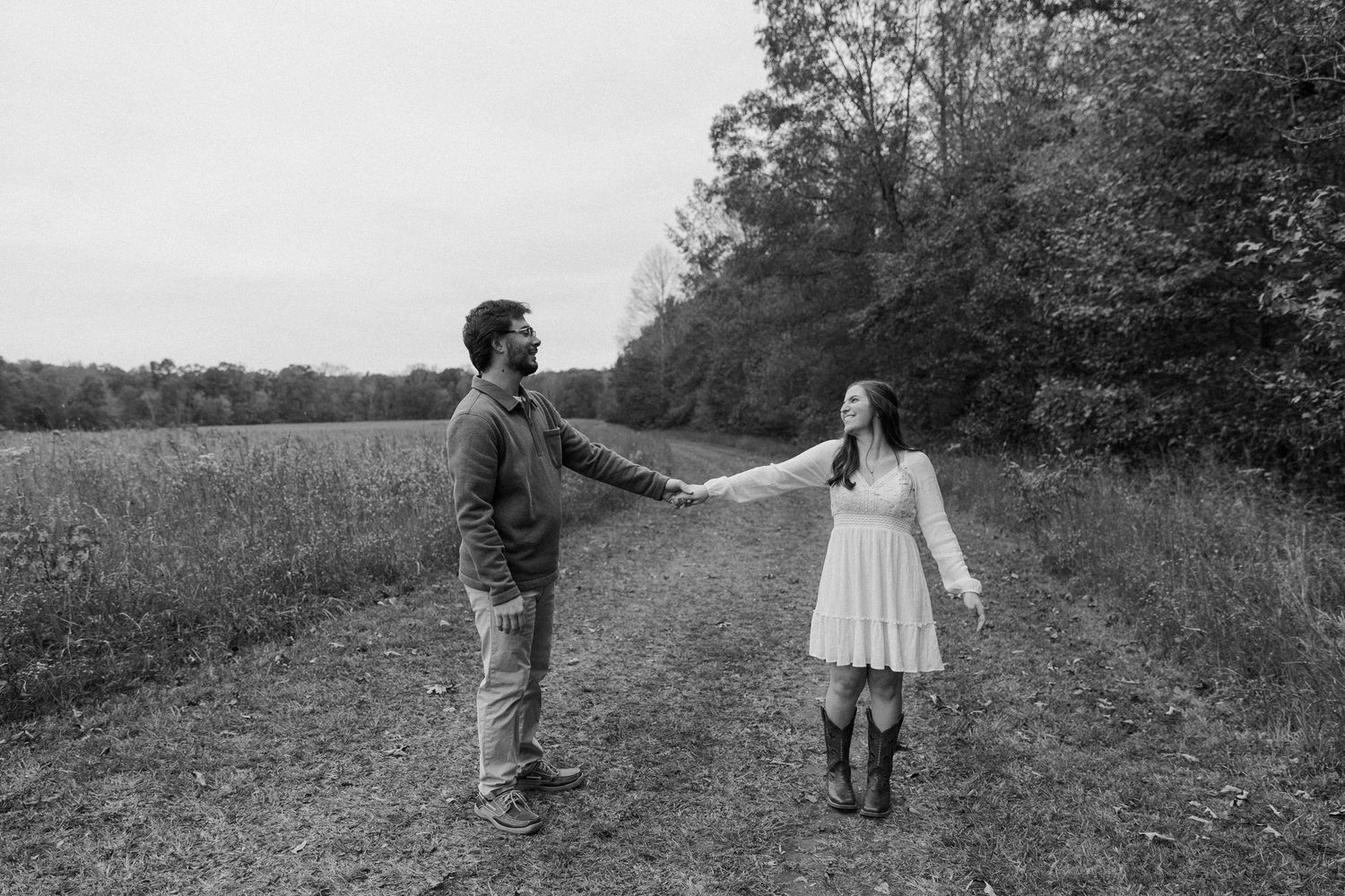 A couple holds hands while standing on a path through a meadow in black and white.