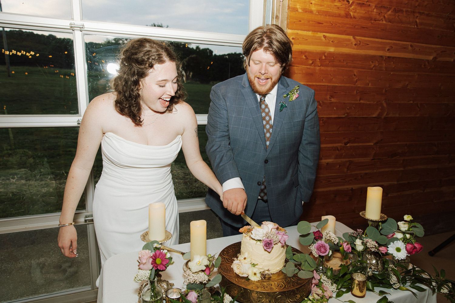 Bride and groom smiling while cutting their wedding cake at a rustic venue decorated with candles and flowers.