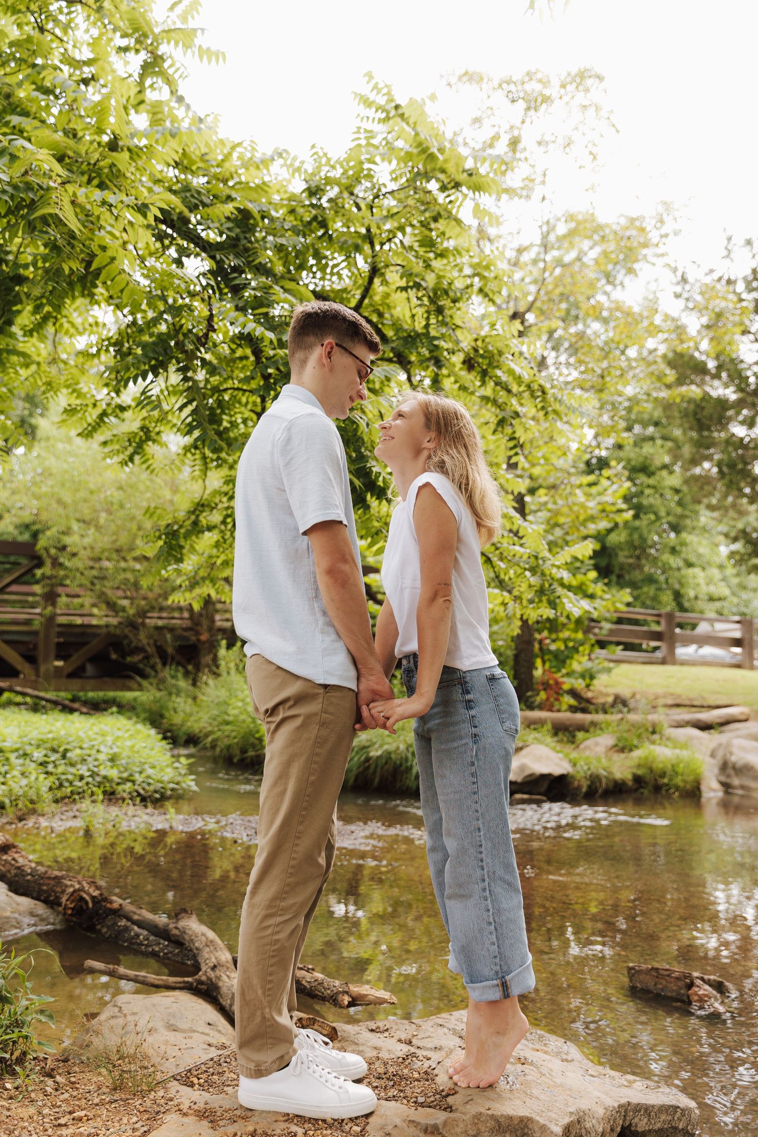 A couple stands together on rocks by a tranquil pond surrounded by lush green trees on a sunny day.