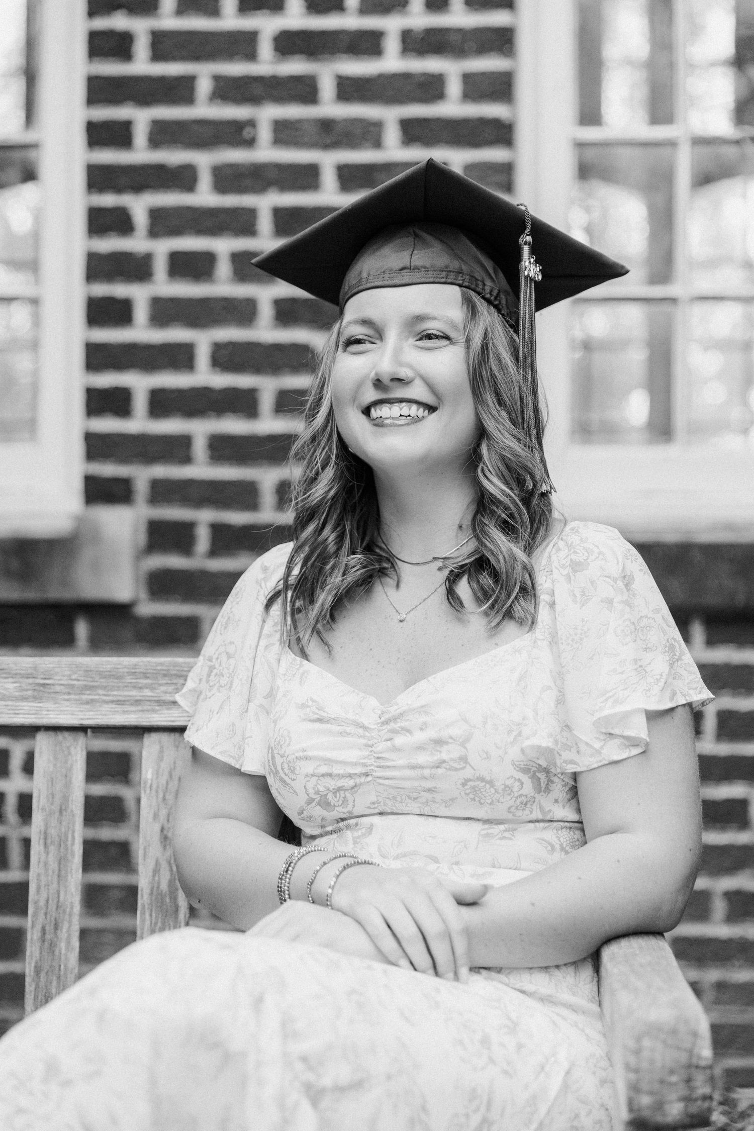 Graduate in white dress and mortarboard cap sits on wooden bench against brick wall, smiling joyfully.