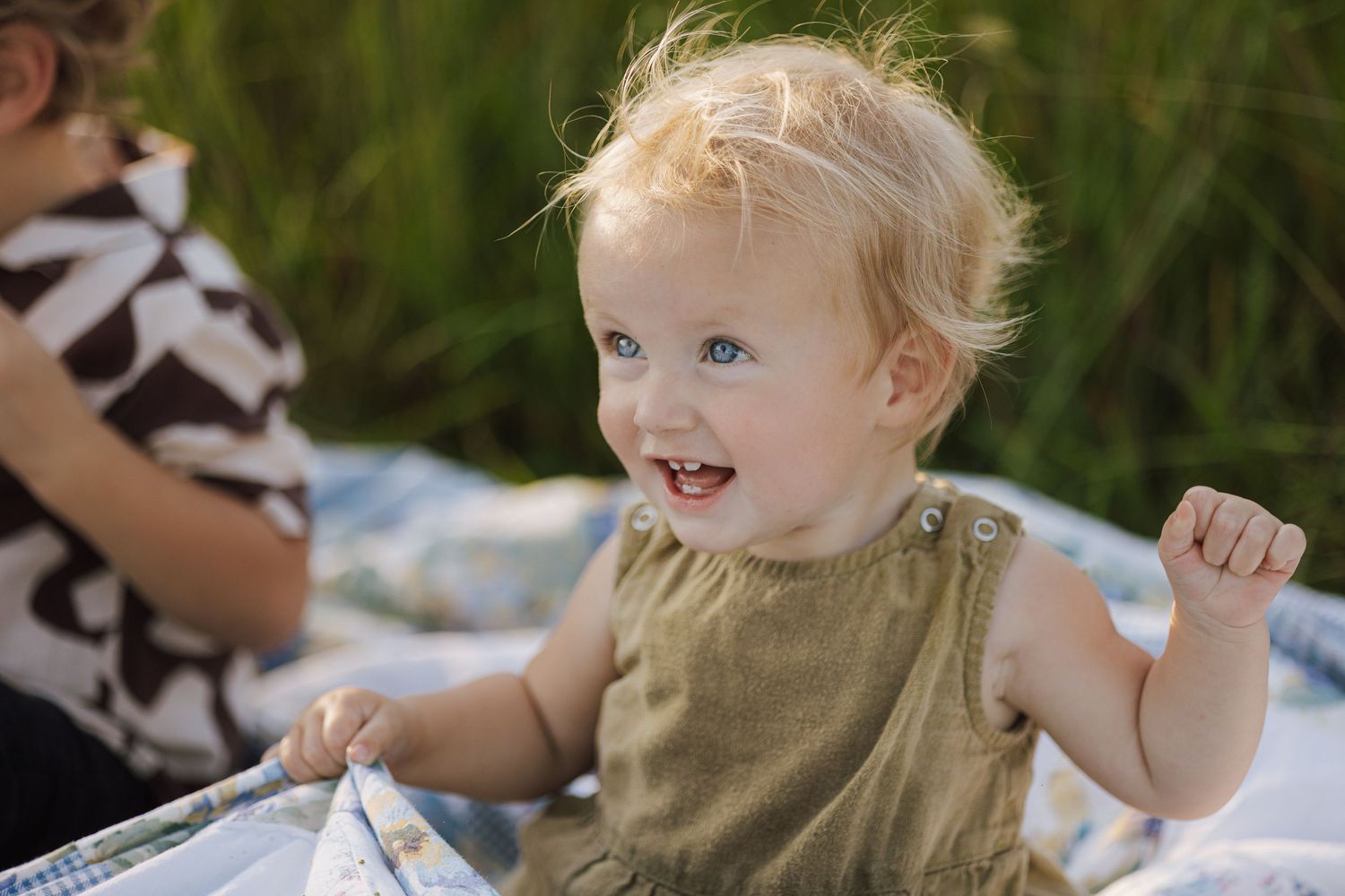 Small child in olive green sleeveless top sits outdoors on white blanket with joyful expression.