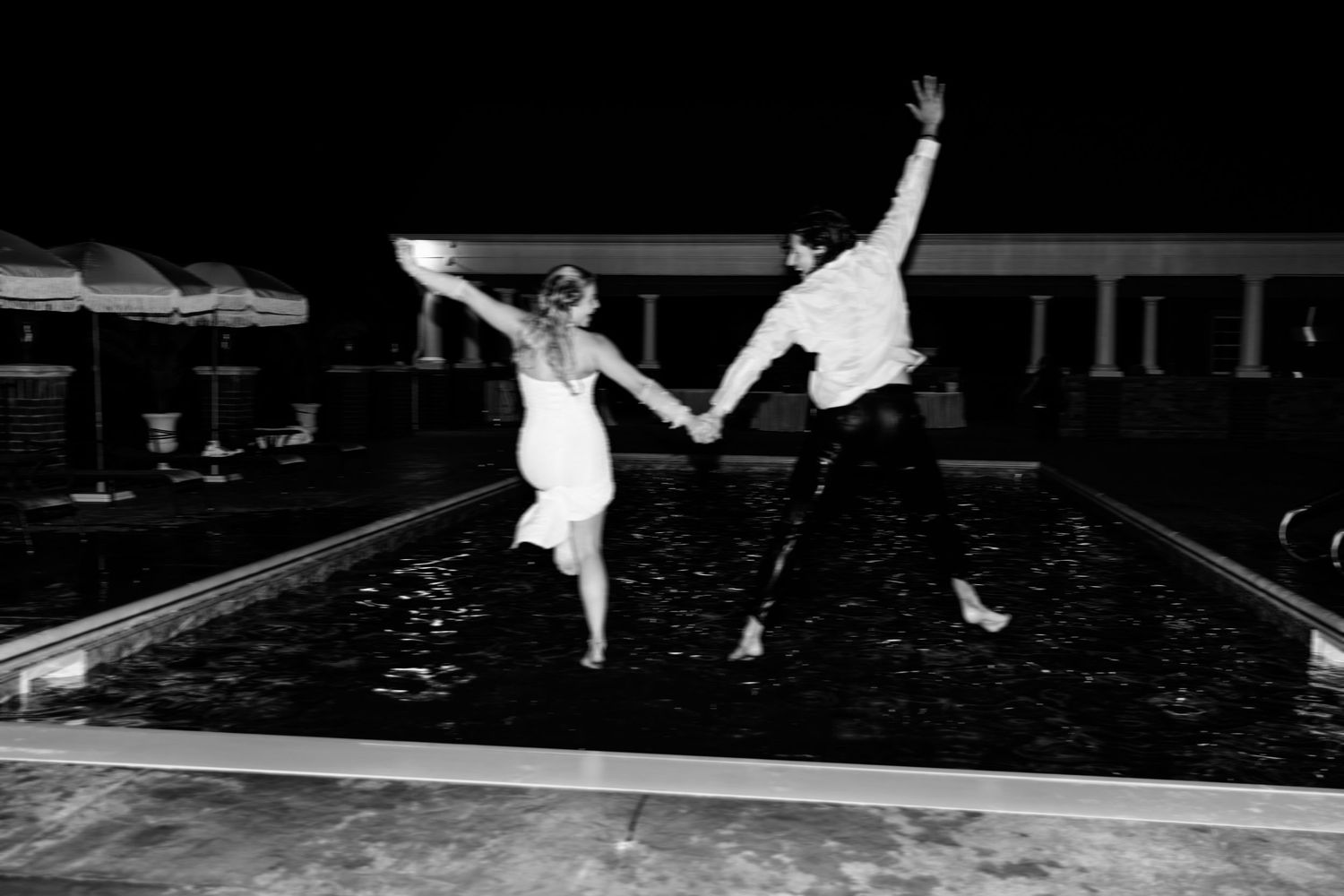 Two people in white clothing jumping playfully by a swimming pool at night.