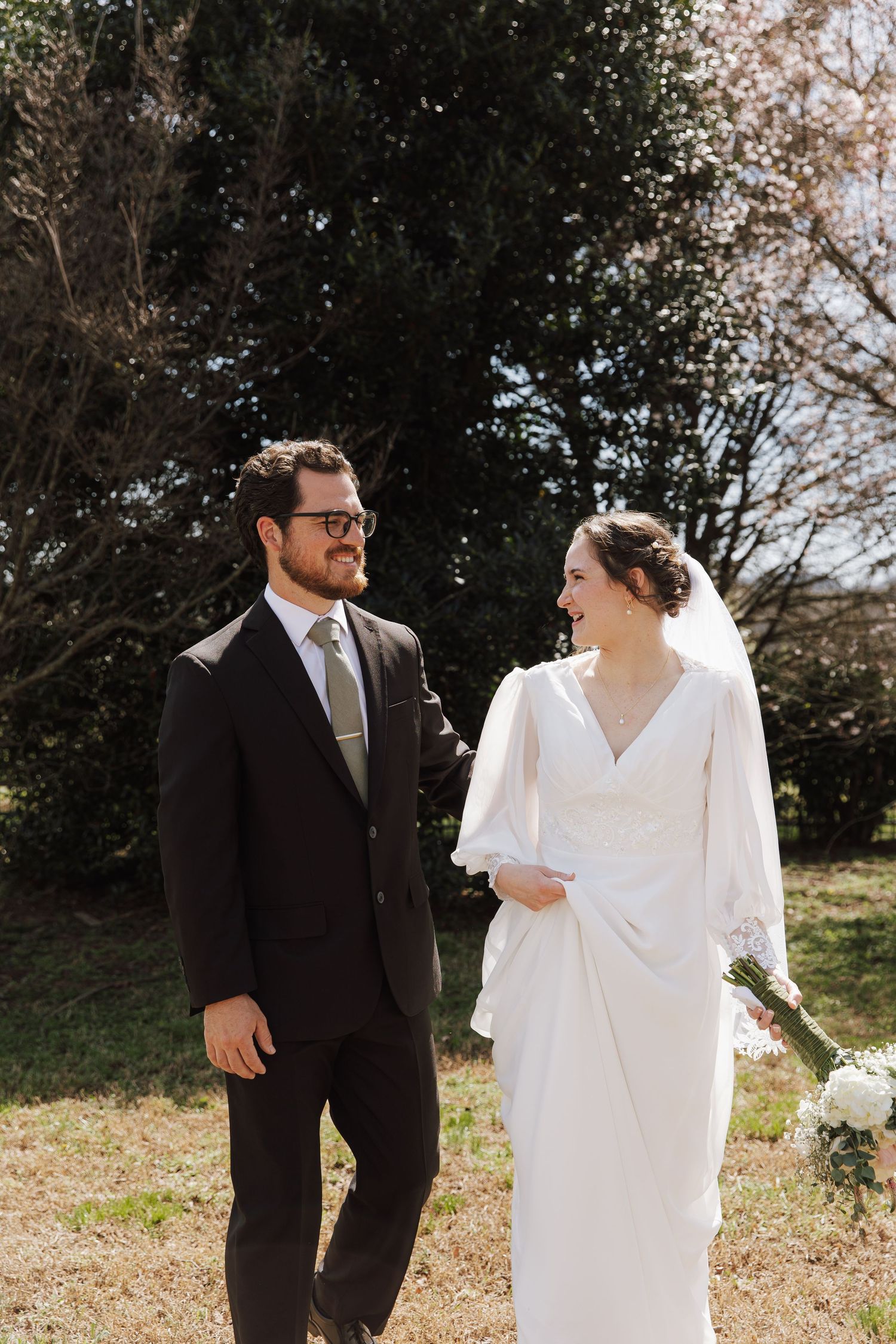 Smiling bride in white gown and groom in dark suit walk together outdoors on wedding day.