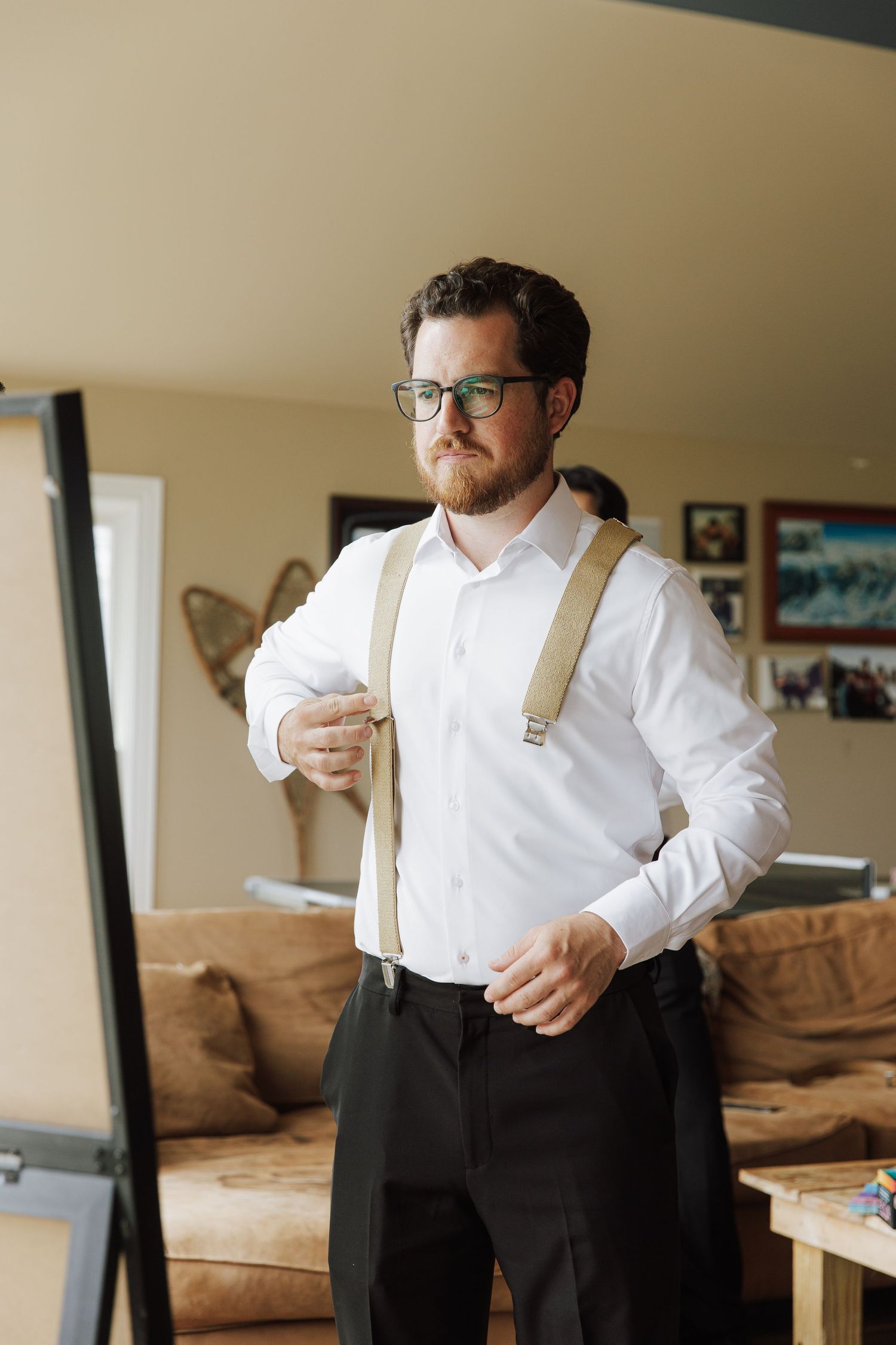 A bearded man in glasses adjusts his tan suspenders over a white dress shirt with black trousers indoors.