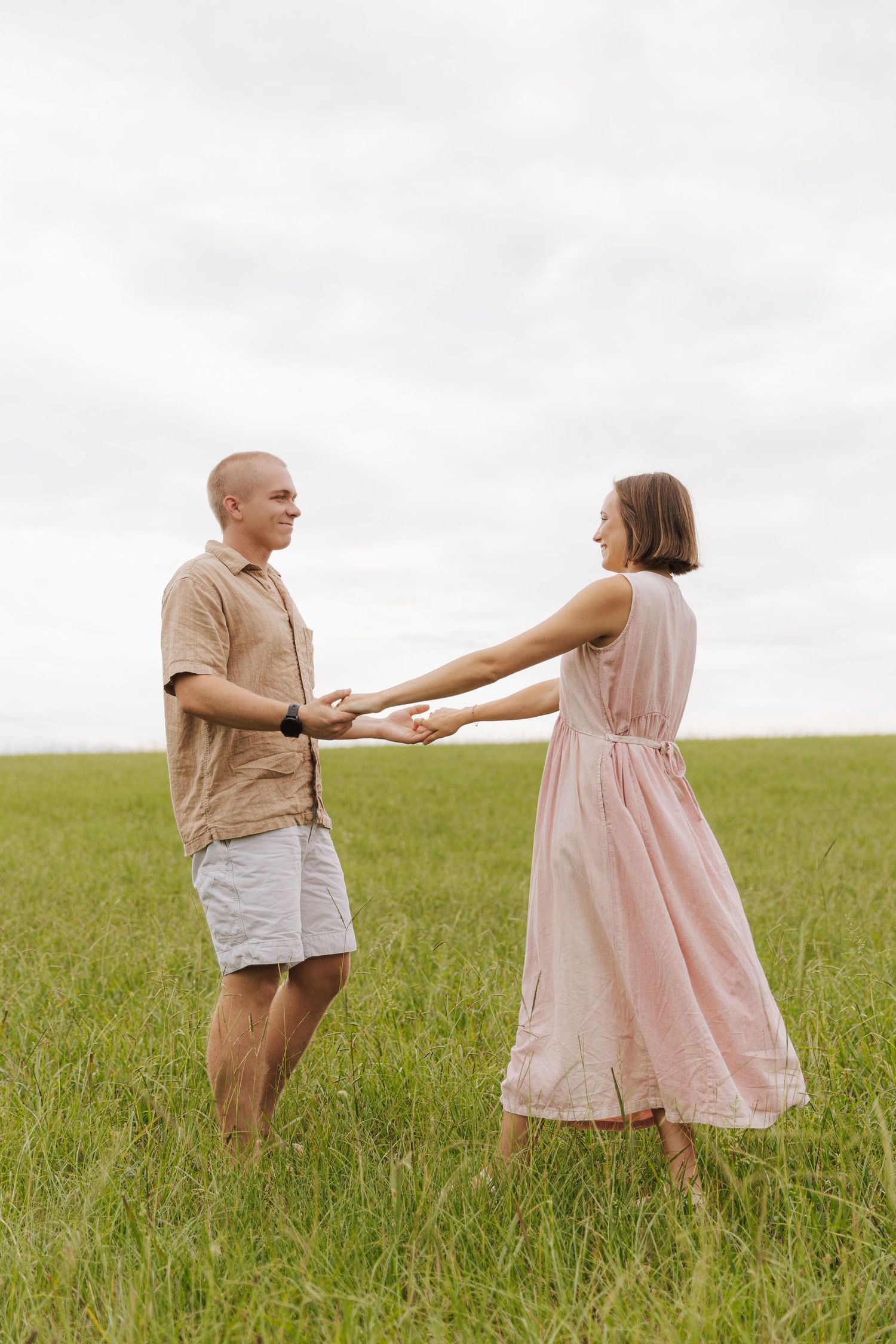 A playful couple holds hands while standing in tall grass on a cloudy day.