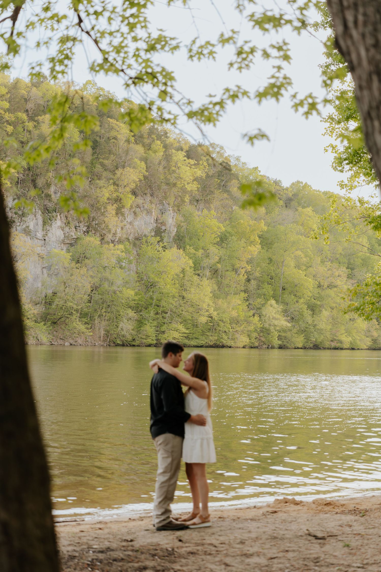 Two people share a romantic moment by a serene lake surrounded by lush green trees on a sunny day.