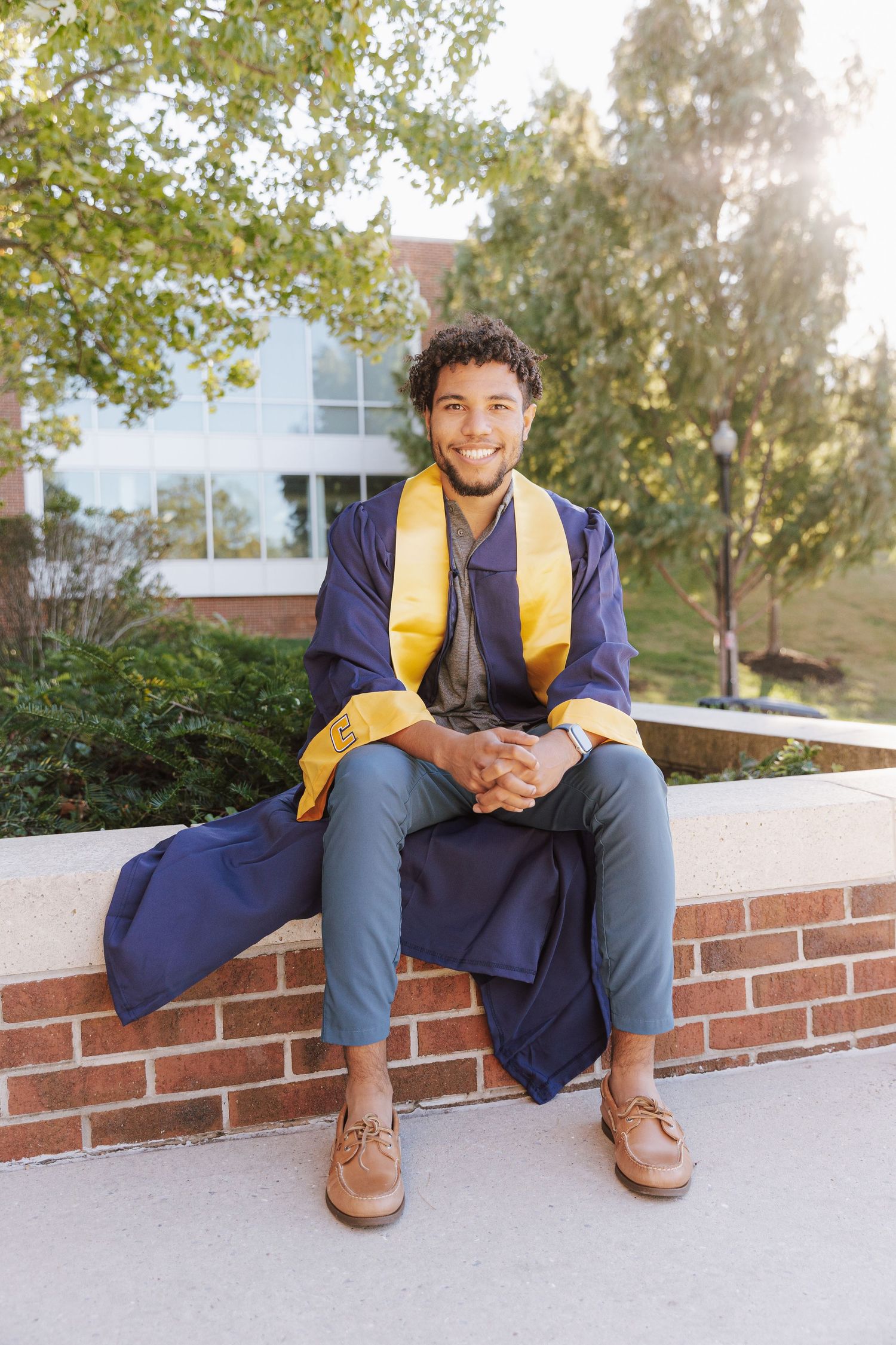 A person wearing a navy varsity jacket and jeans sits casually on a brick ledge outdoors near trees and buildings.