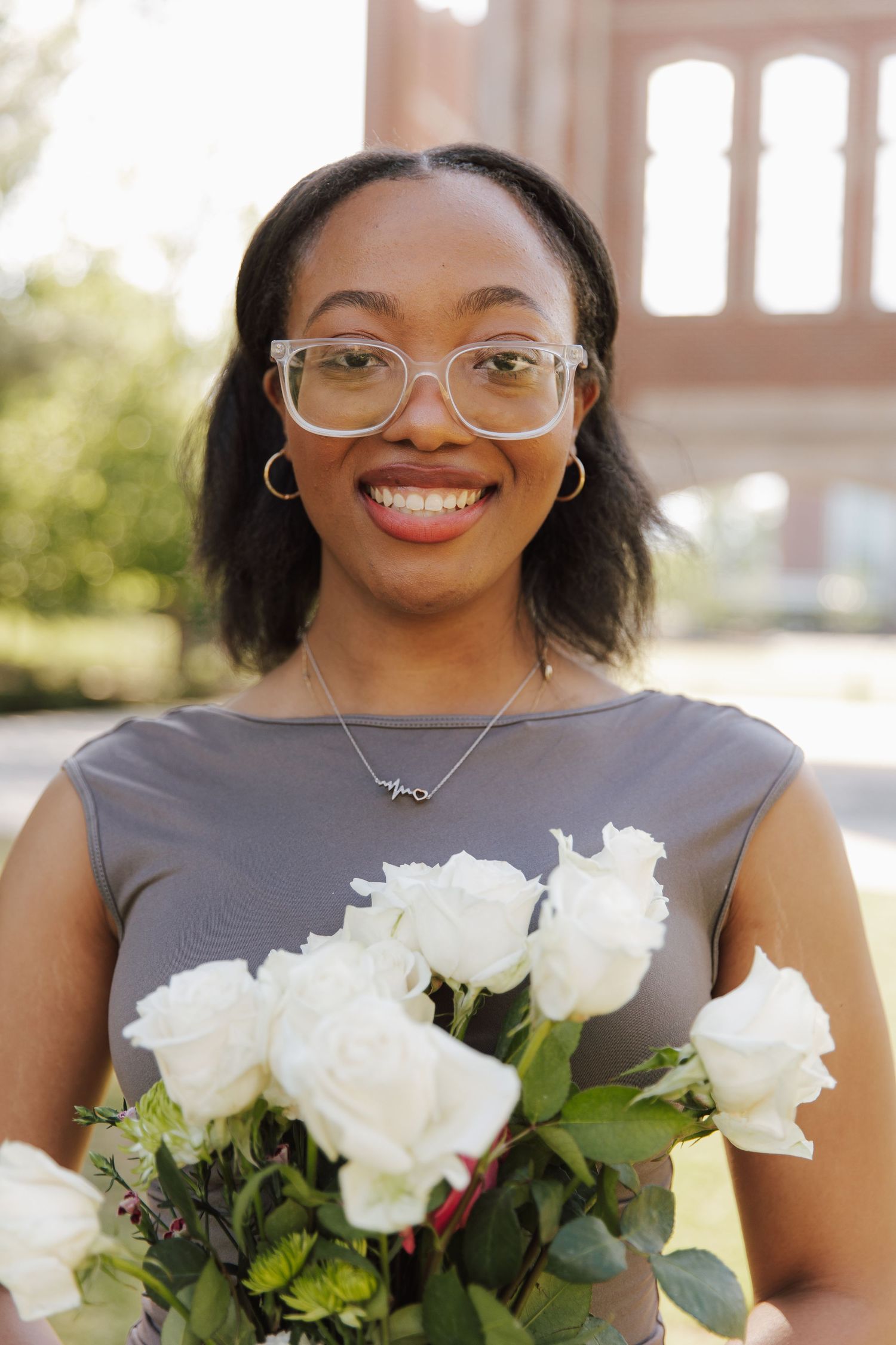 Smiling young woman in gray dress holding white roses bouquet outdoors.