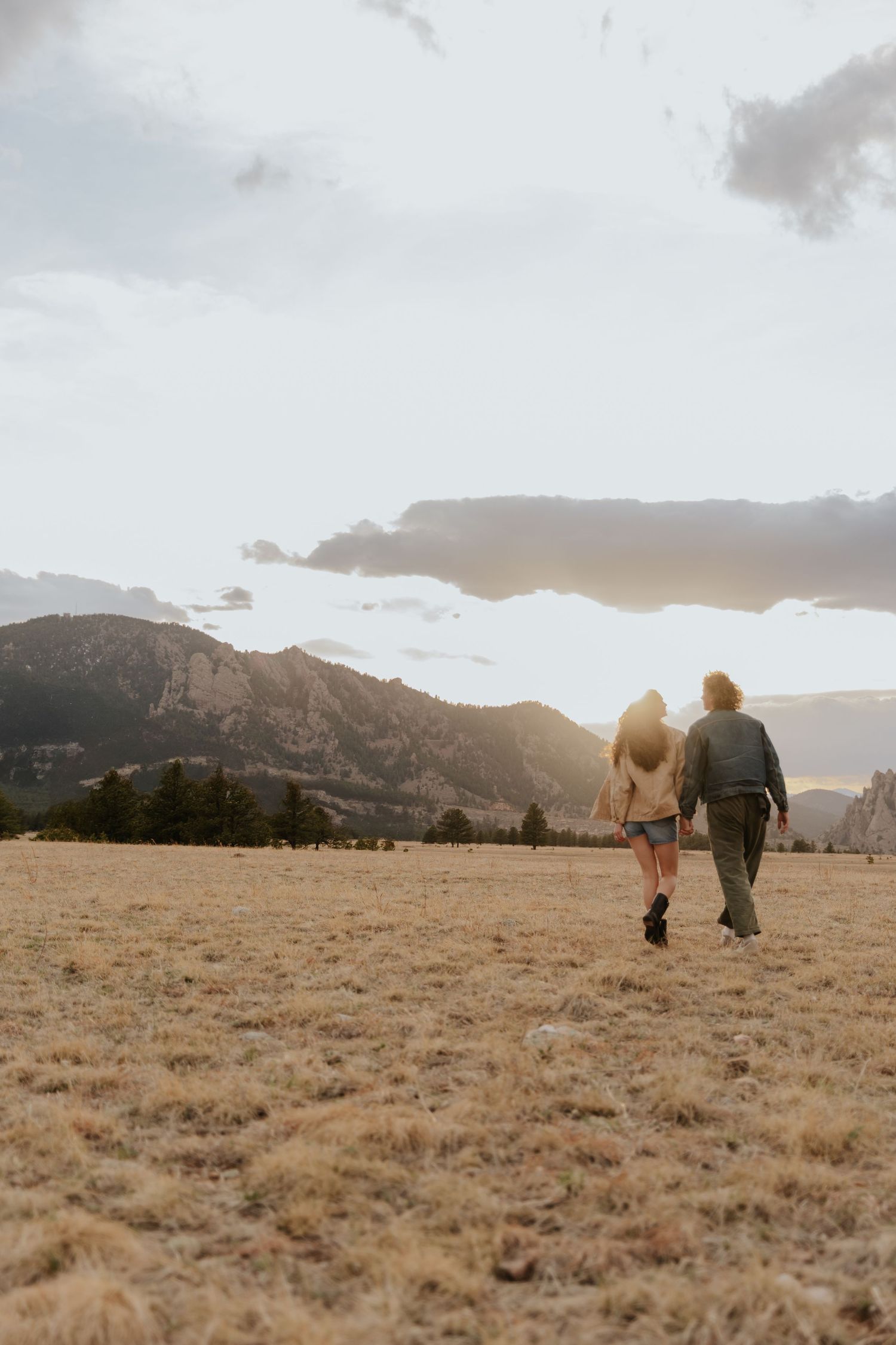 Two people walk together through a grassy field at sunset with mountains in the background.