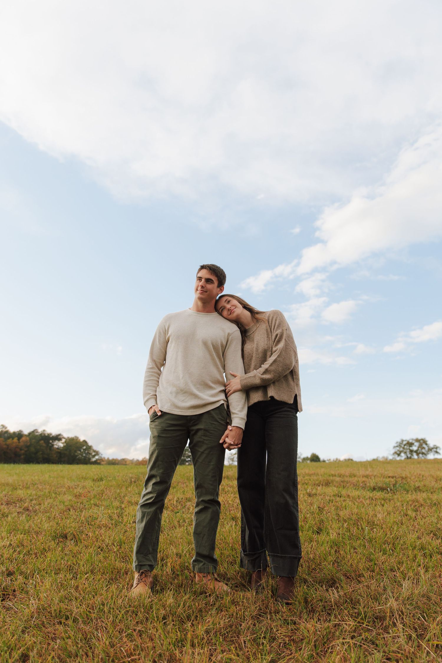 Couple in casual sweaters embrace while standing in a sunlit meadow.