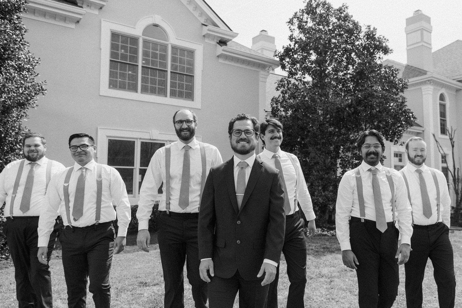 Black and white photo of groom and groomsmen in dress shirts and ties posing outside elegant venue.